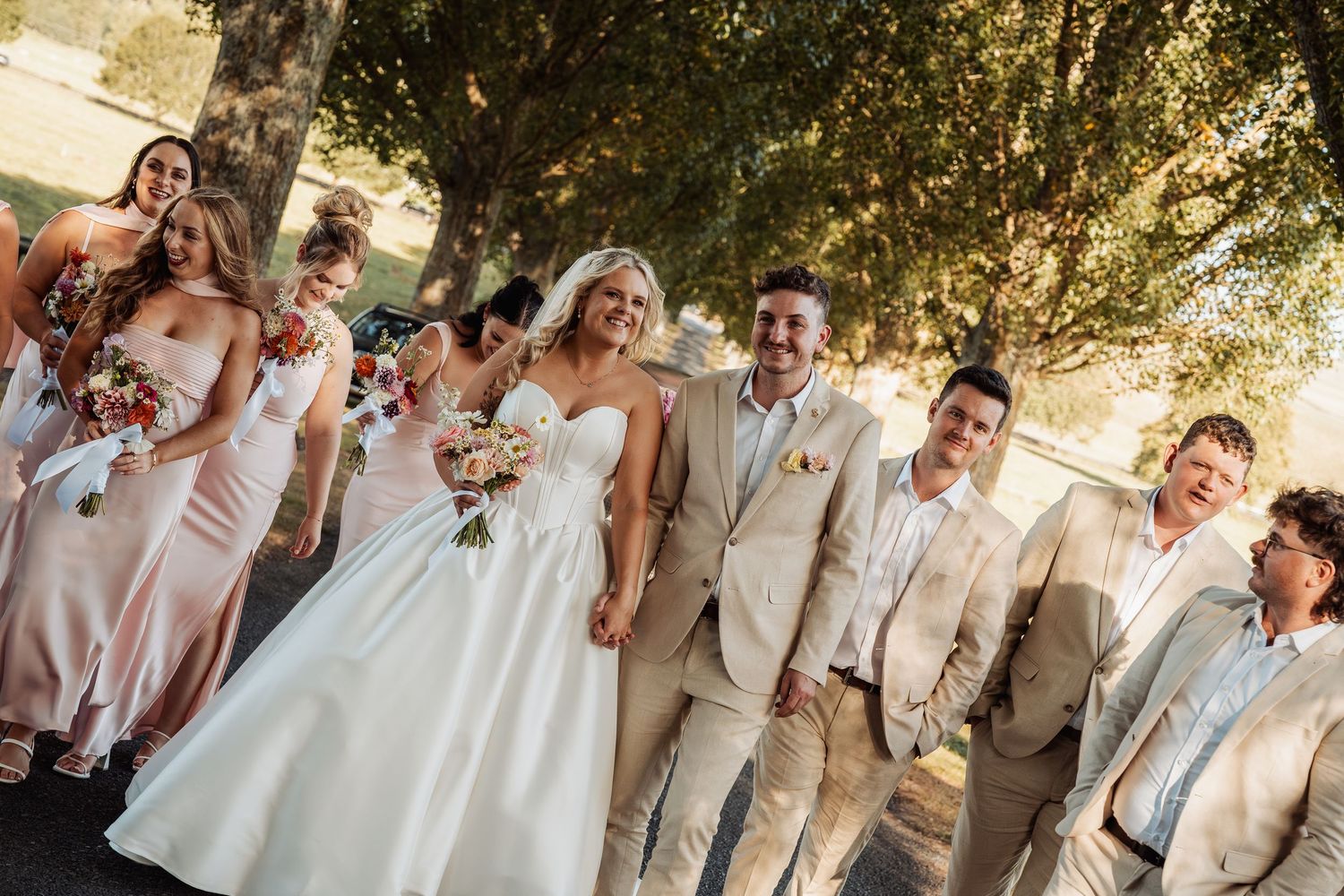 Wedding party in beige and champagne attire laughing together under shaded trees.
