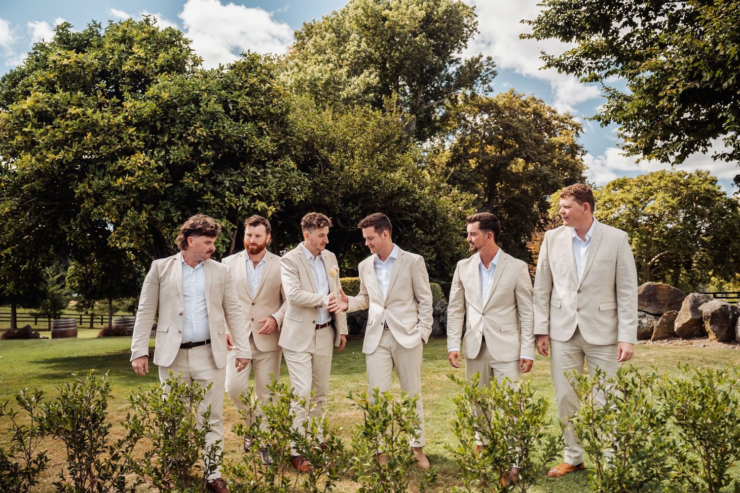 Six groomsmen in beige suits walking together through a sunny garden setting.