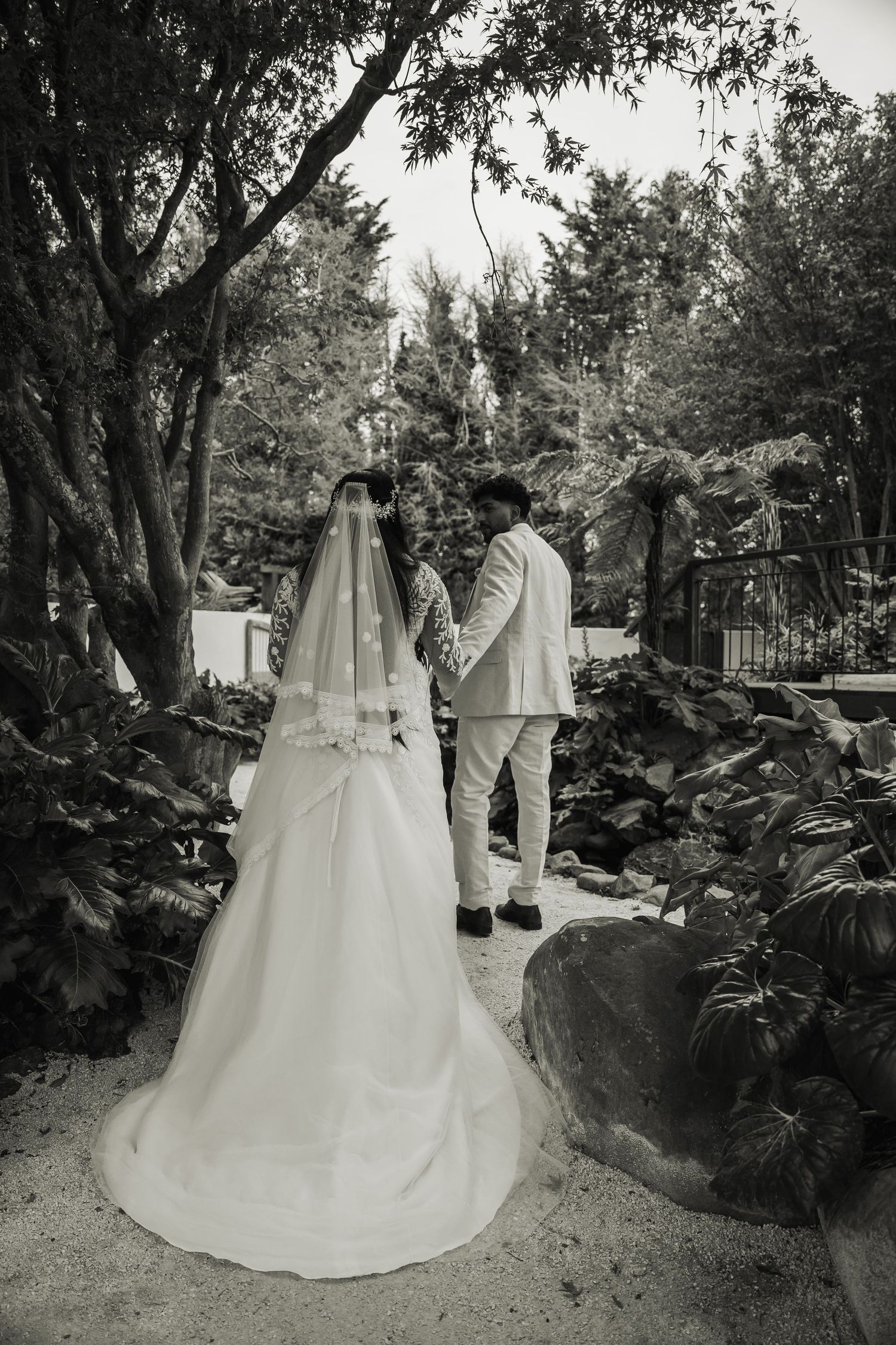 Black and white photo of a bride and groom holding hands walking through a lush garden on their wedding day.