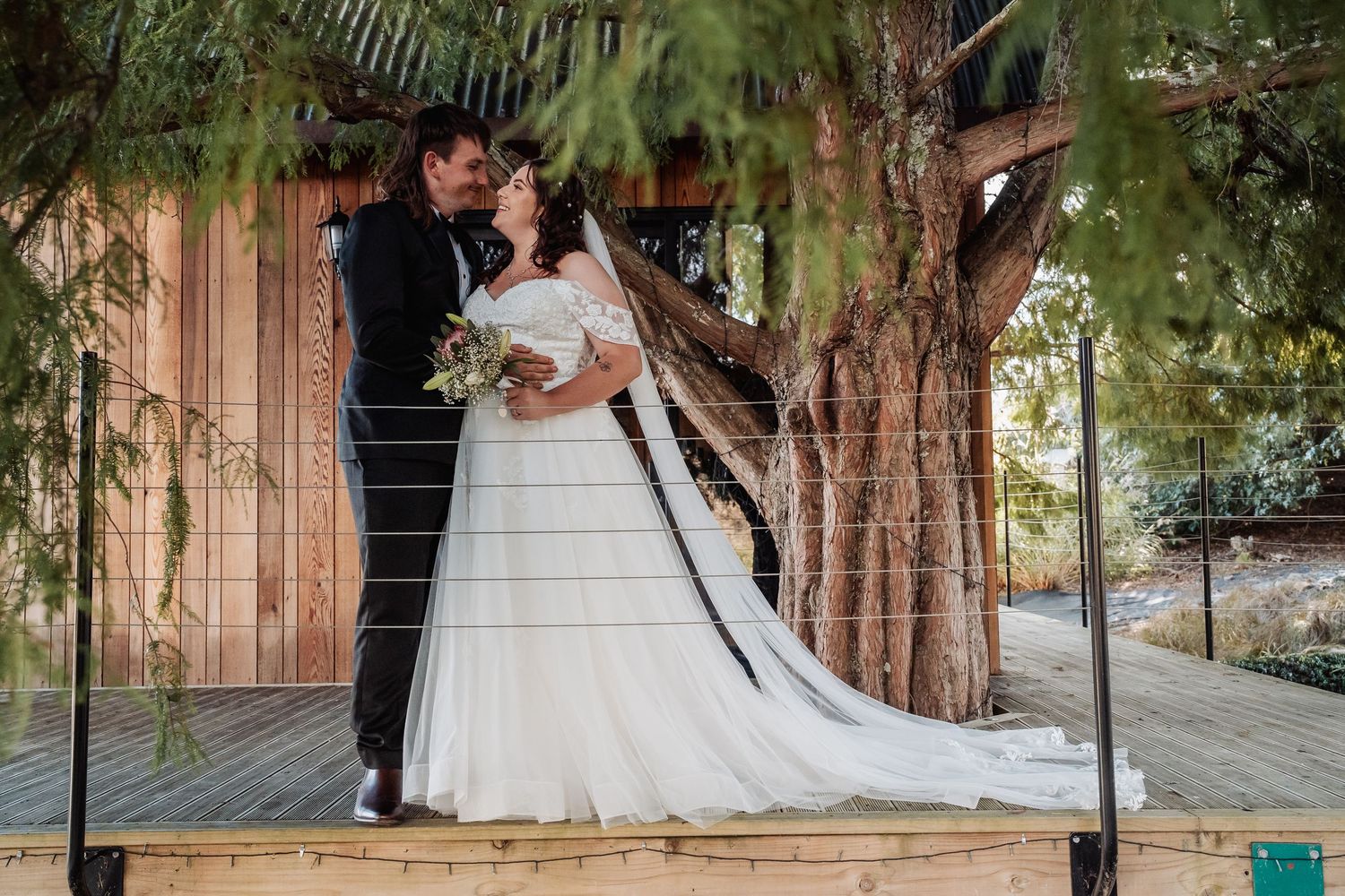 Bride and groom share a tender moment under tree canopy at rustic outdoor wedding venue.
