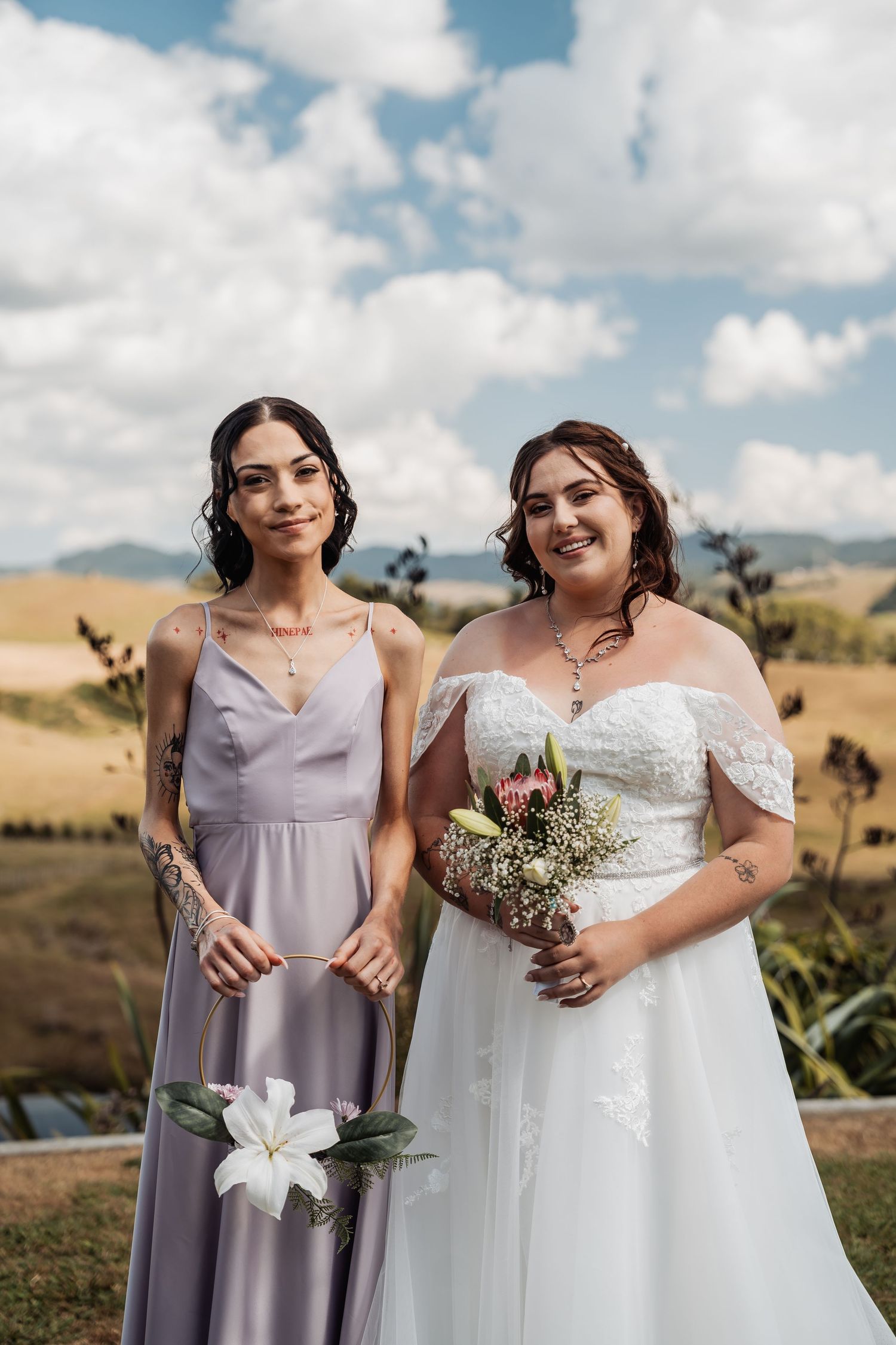 Bride in white off-shoulder gown and bridesmaid in lavender dress holding flowers outdoors with scenic hills backdrop.