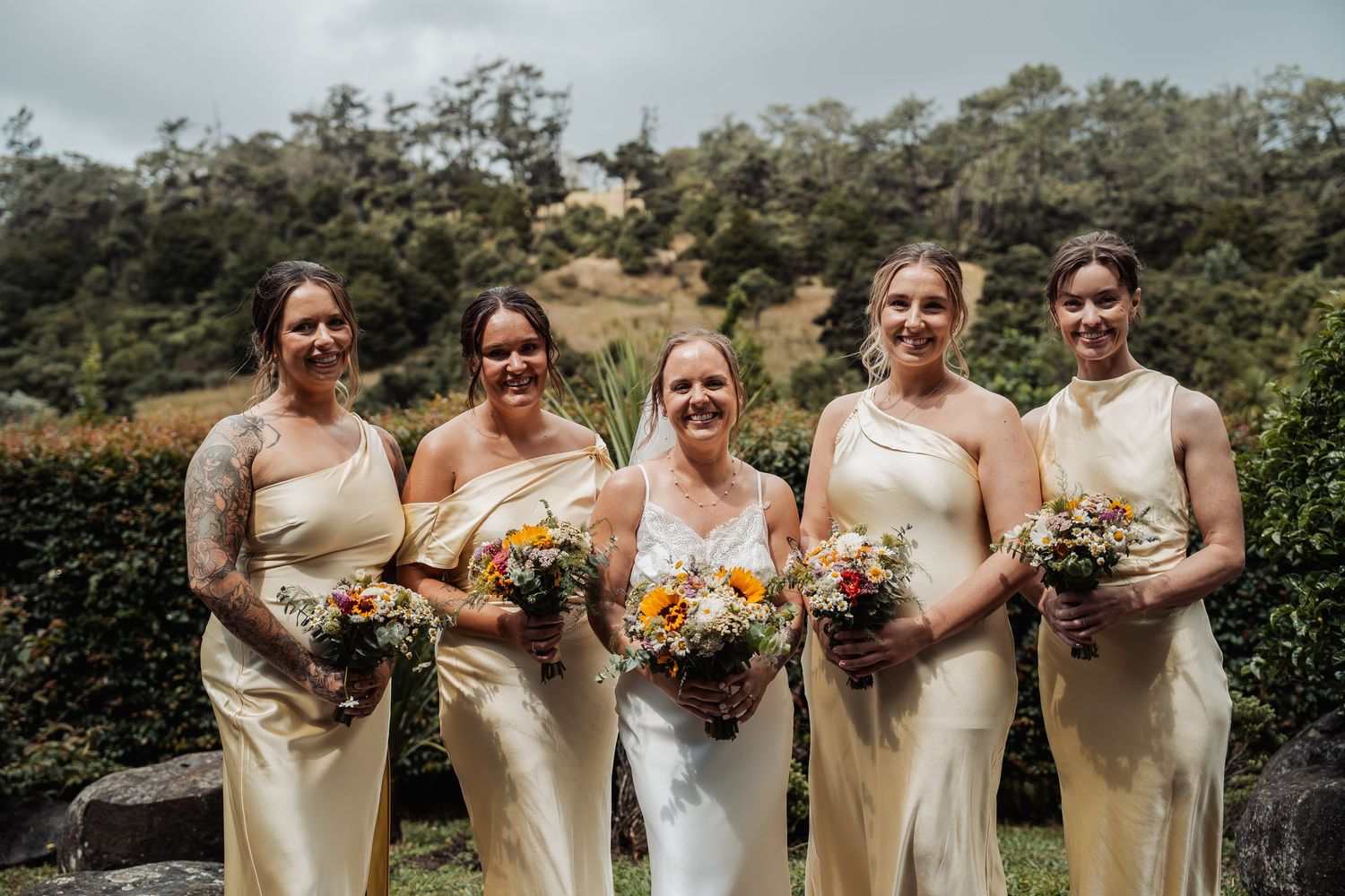 Bride with four bridesmaids in matching champagne satin dresses holding colorful bouquets outdoors.