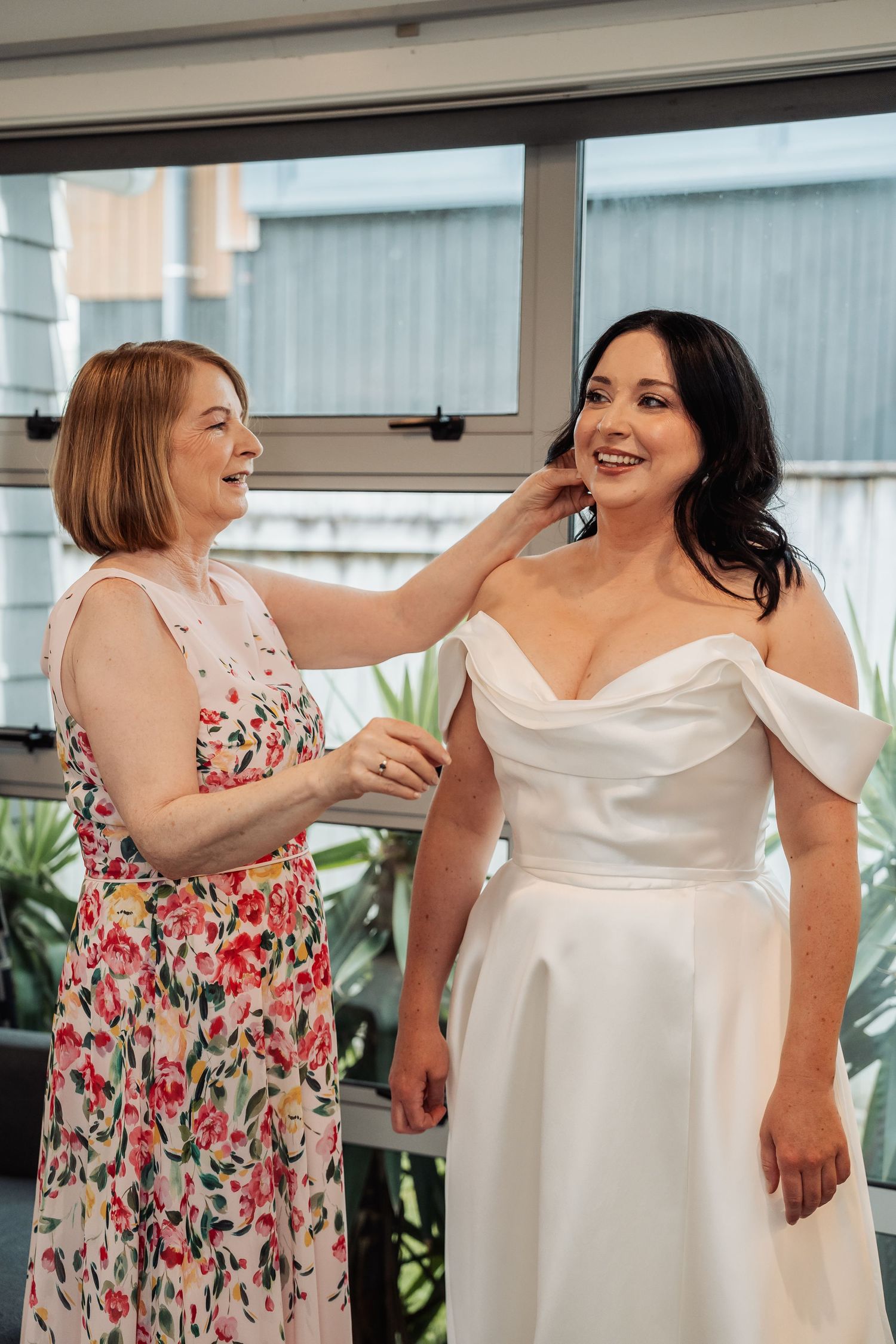 A woman in a floral dress adjusts the hair of a smiling bride in an off-shoulder white wedding gown.