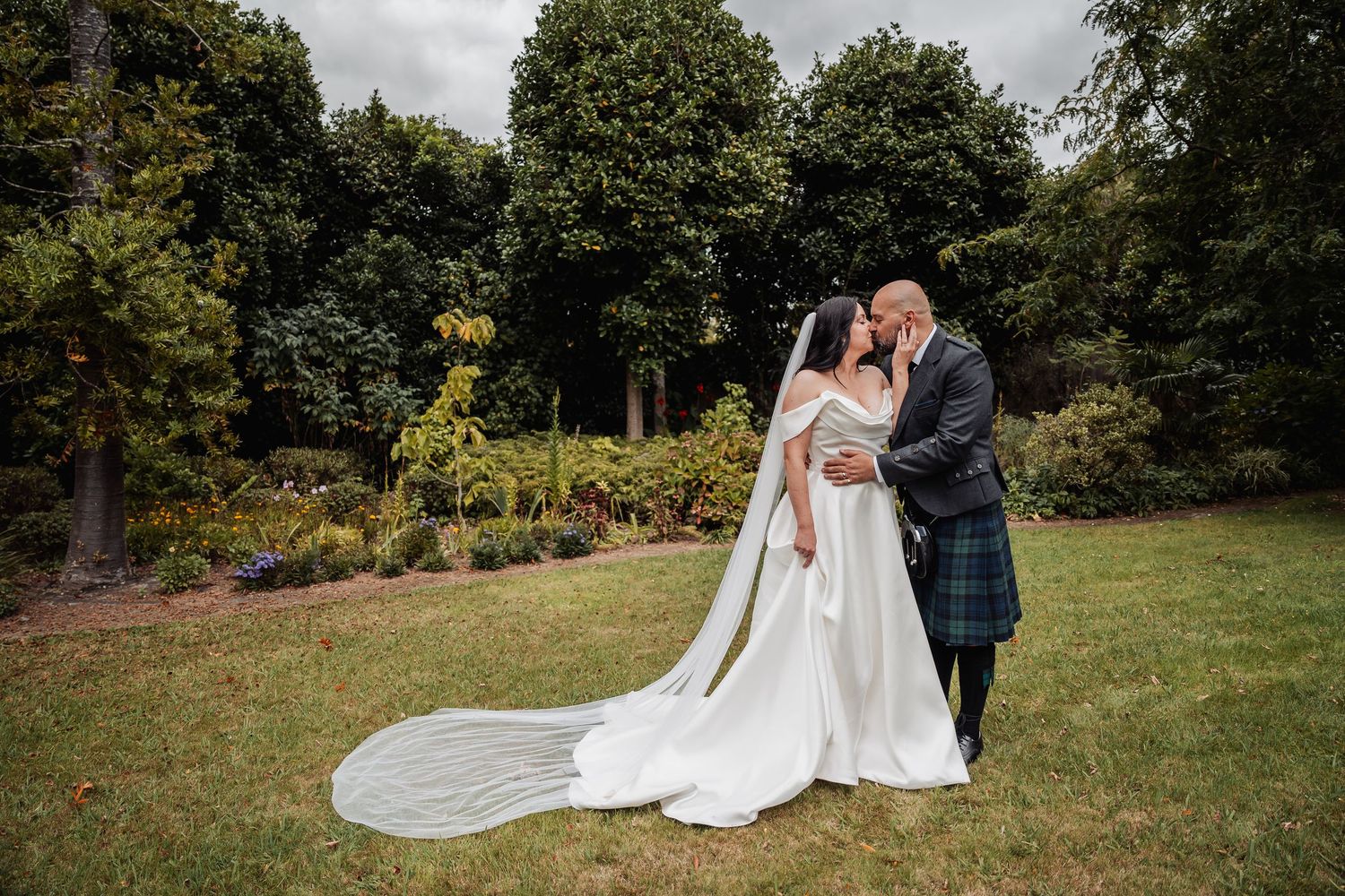 Newlyweds share a kiss in lush garden, bride wearing long veil and off-shoulder gown.