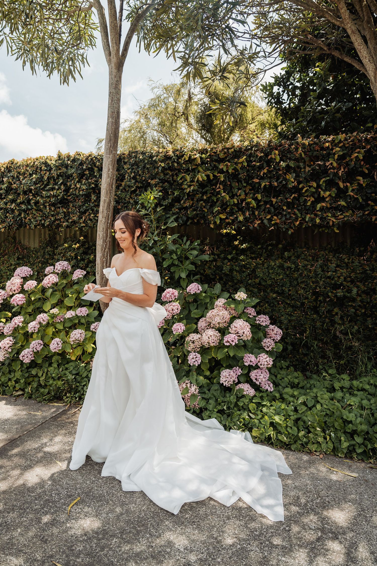 Bride in off-shoulder white gown with train standing among pink hydrangeas in garden.