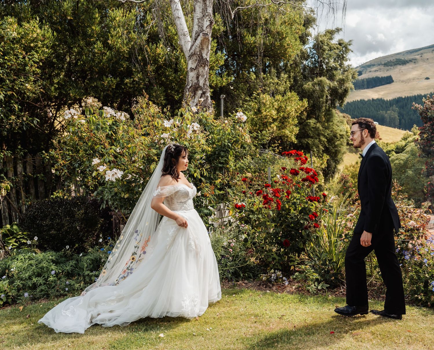 Bride in a white off-shoulder gown with floral veil stands facing groom in a garden with roses and hills.