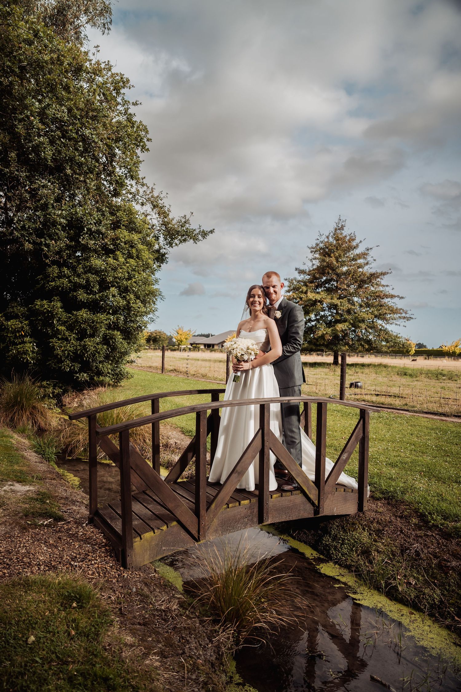 Bride and groom posing together on a rustic wooden bridge surrounded by lush countryside scenery.