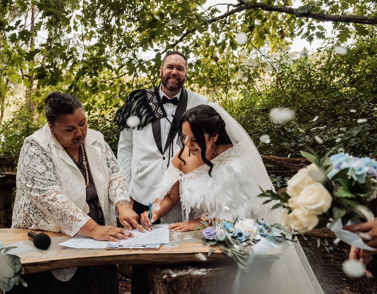 Bride and groom sign wedding documents with officiant in an outdoor garden setting.