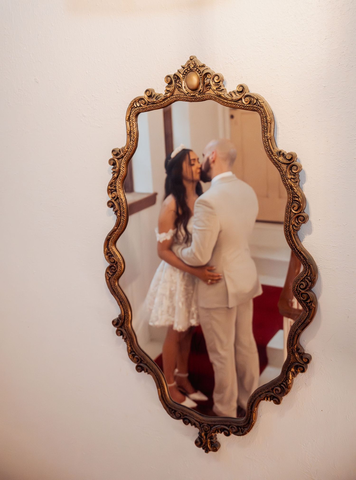 Couple sharing a kiss reflected in ornate gold vintage mirror mounted on white wall.