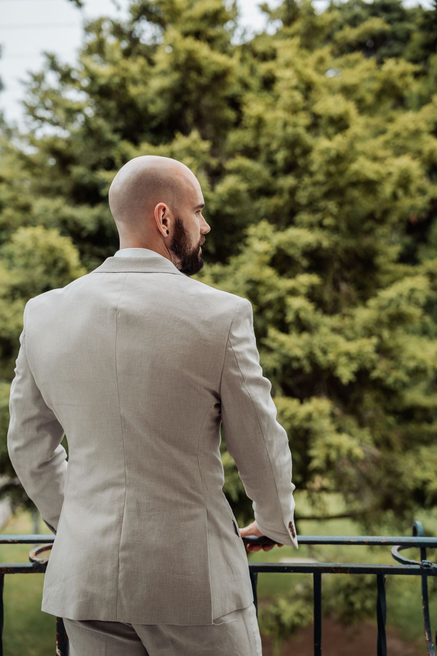 Bald groom in beige suit stands at outdoor railing surrounded by lush green trees.