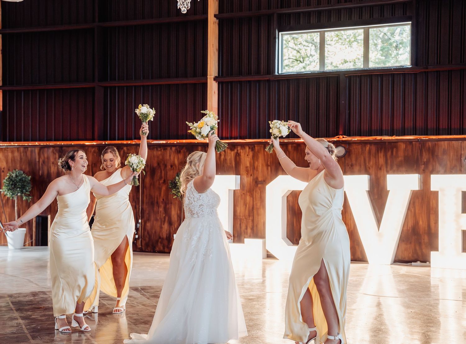 Bride and bridesmaids in white dresses raise bouquets inside barn with LOVE sign.
