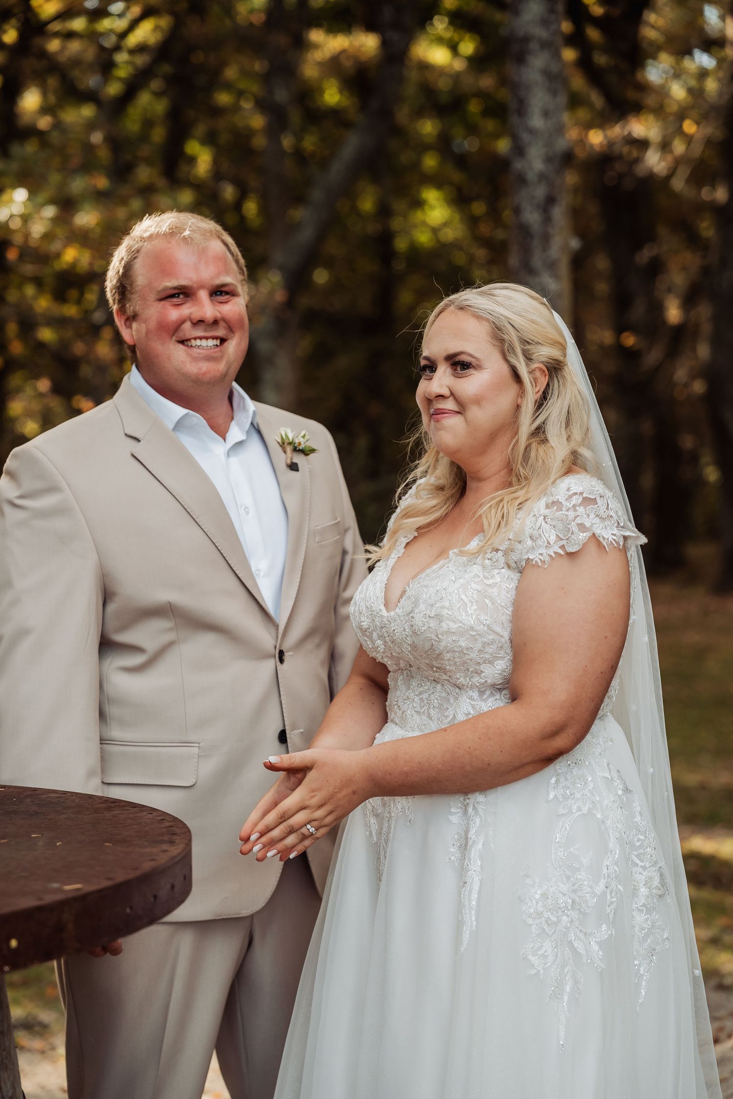 Smiling bride in lace gown and groom in tan suit stand together outdoors at a woodland wedding ceremony.