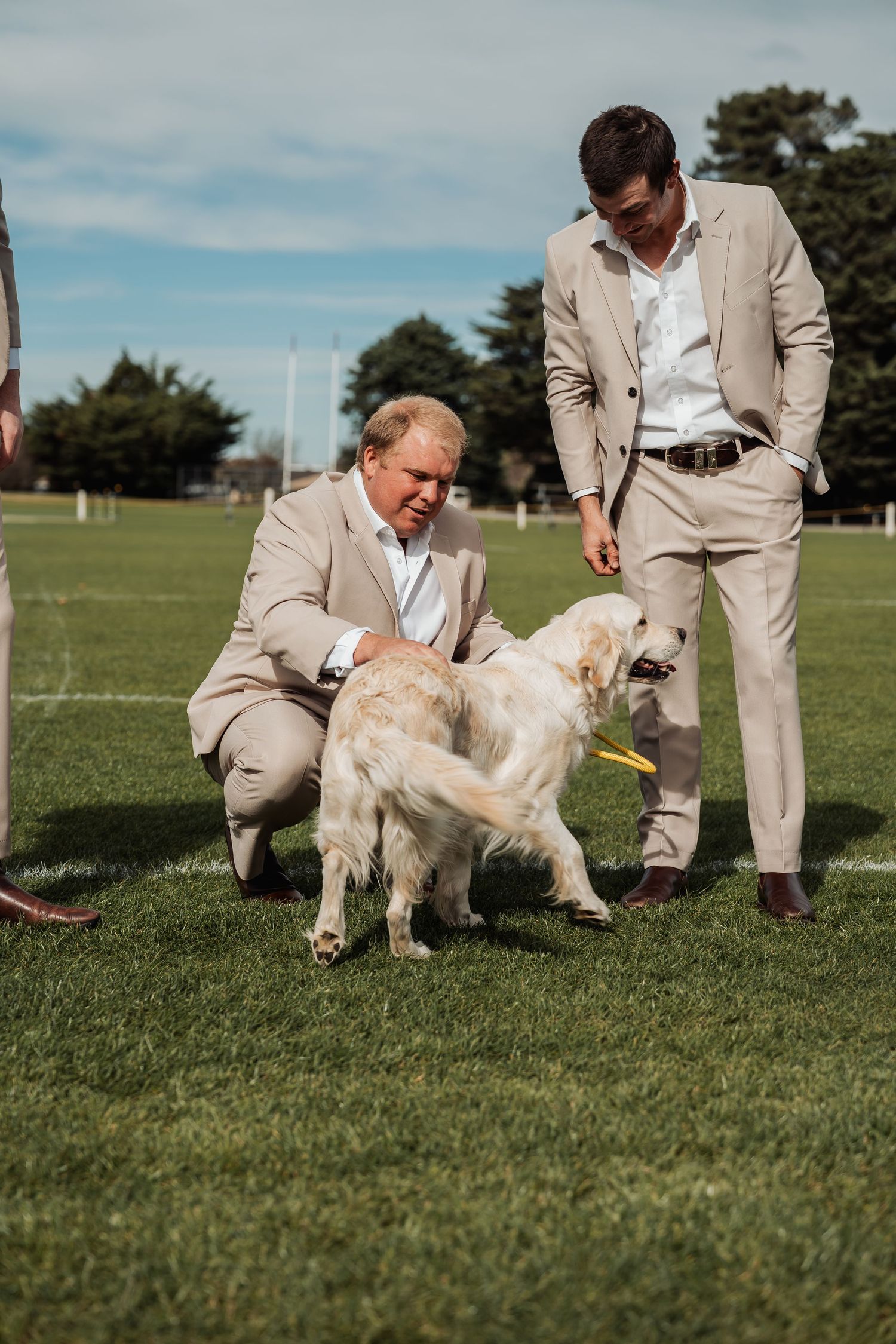 Two men in beige suits interact with a white dog on a green sports field outdoors.