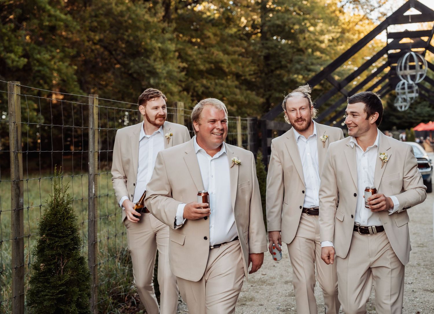 Four groomsmen in matching tan suits laughing and walking together at an outdoor wedding venue.