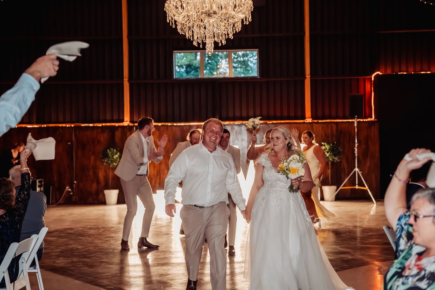 Bride and groom make a grand entrance into a rustic barn reception hall as guests cheer and celebrate.