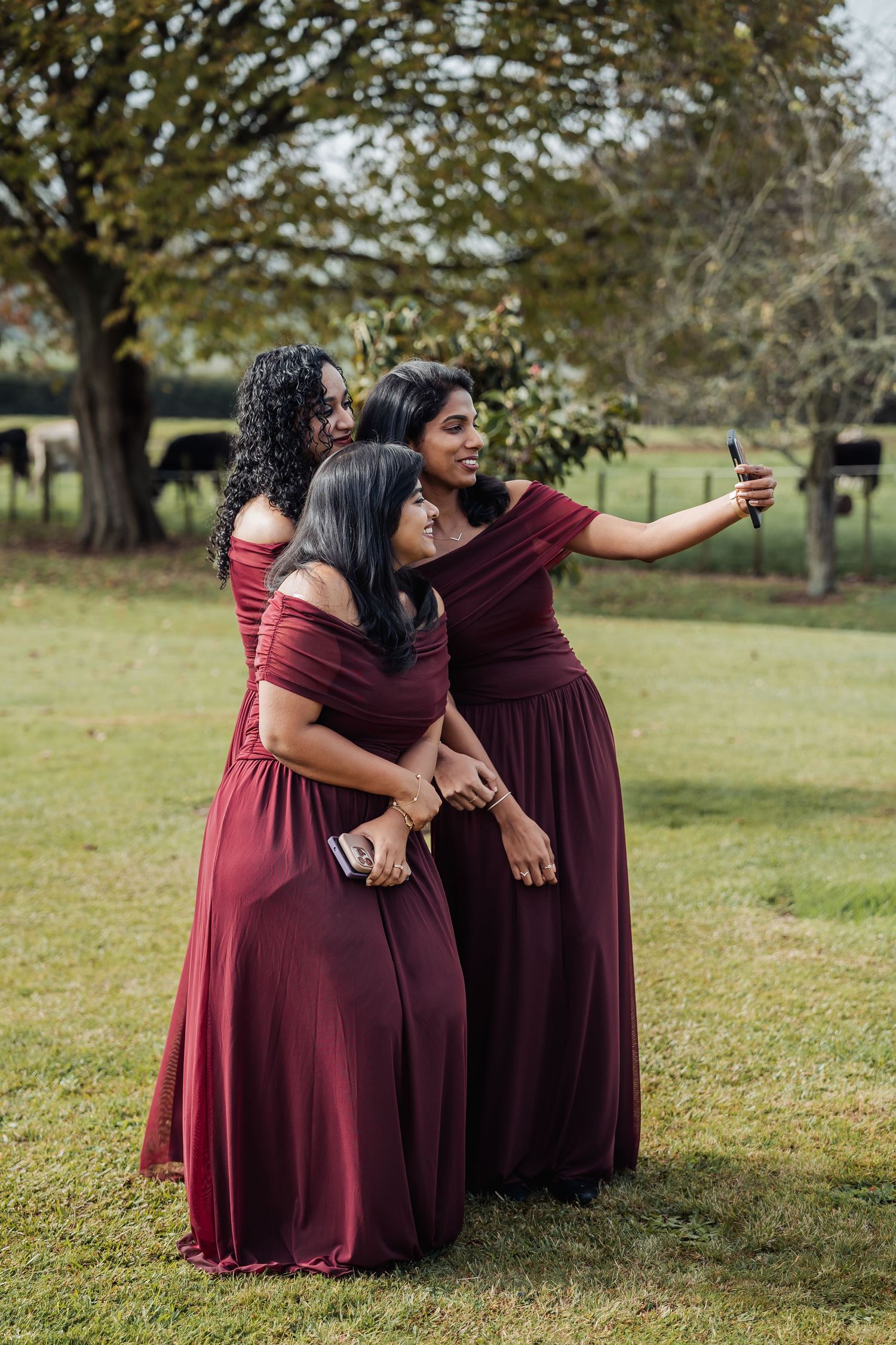 Three bridesmaids in burgundy gowns take a selfie together on green lawn at outdoor wedding.