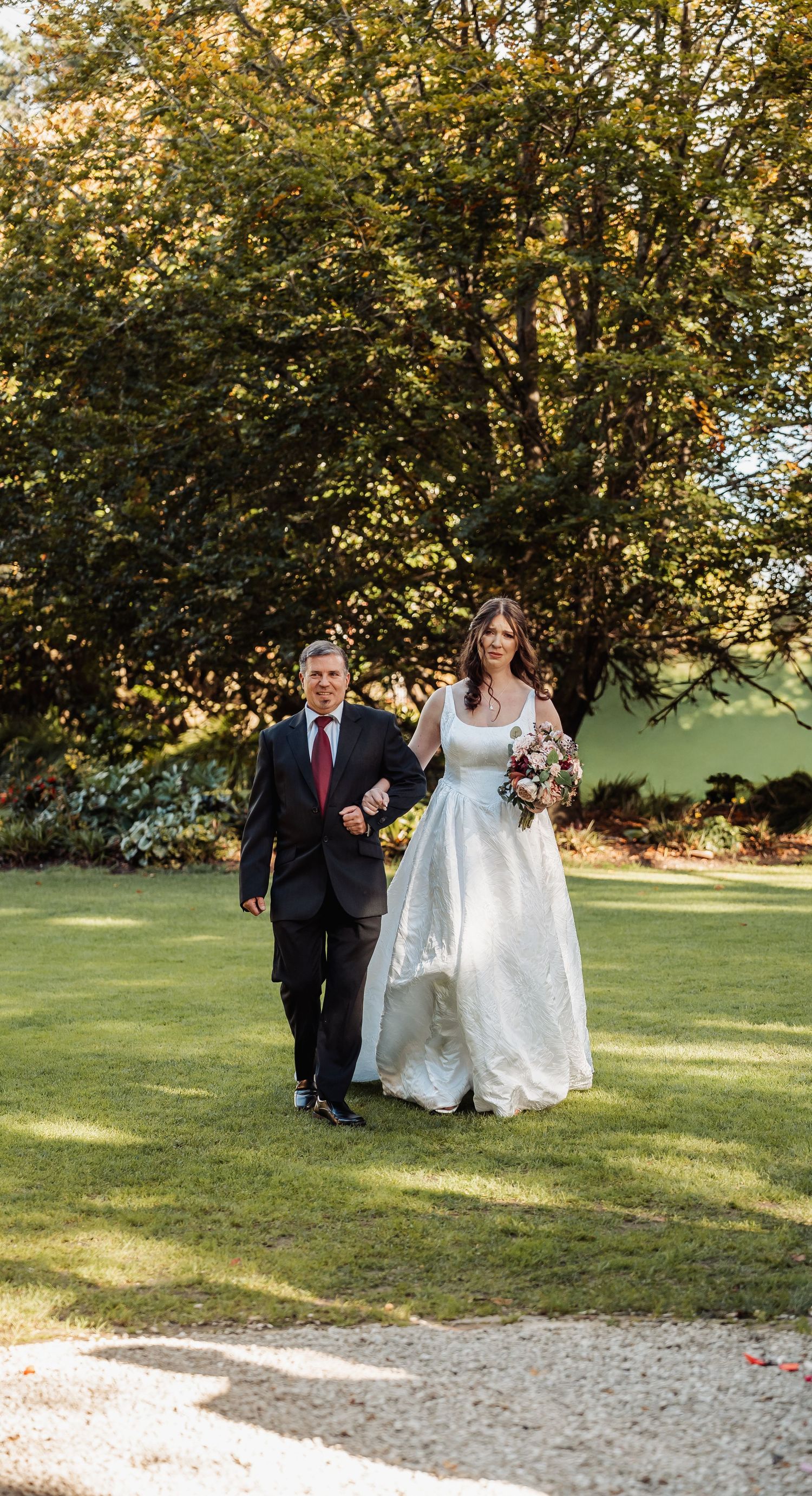 Bride in white gown walks arm in arm with suited man across sunny green lawn.