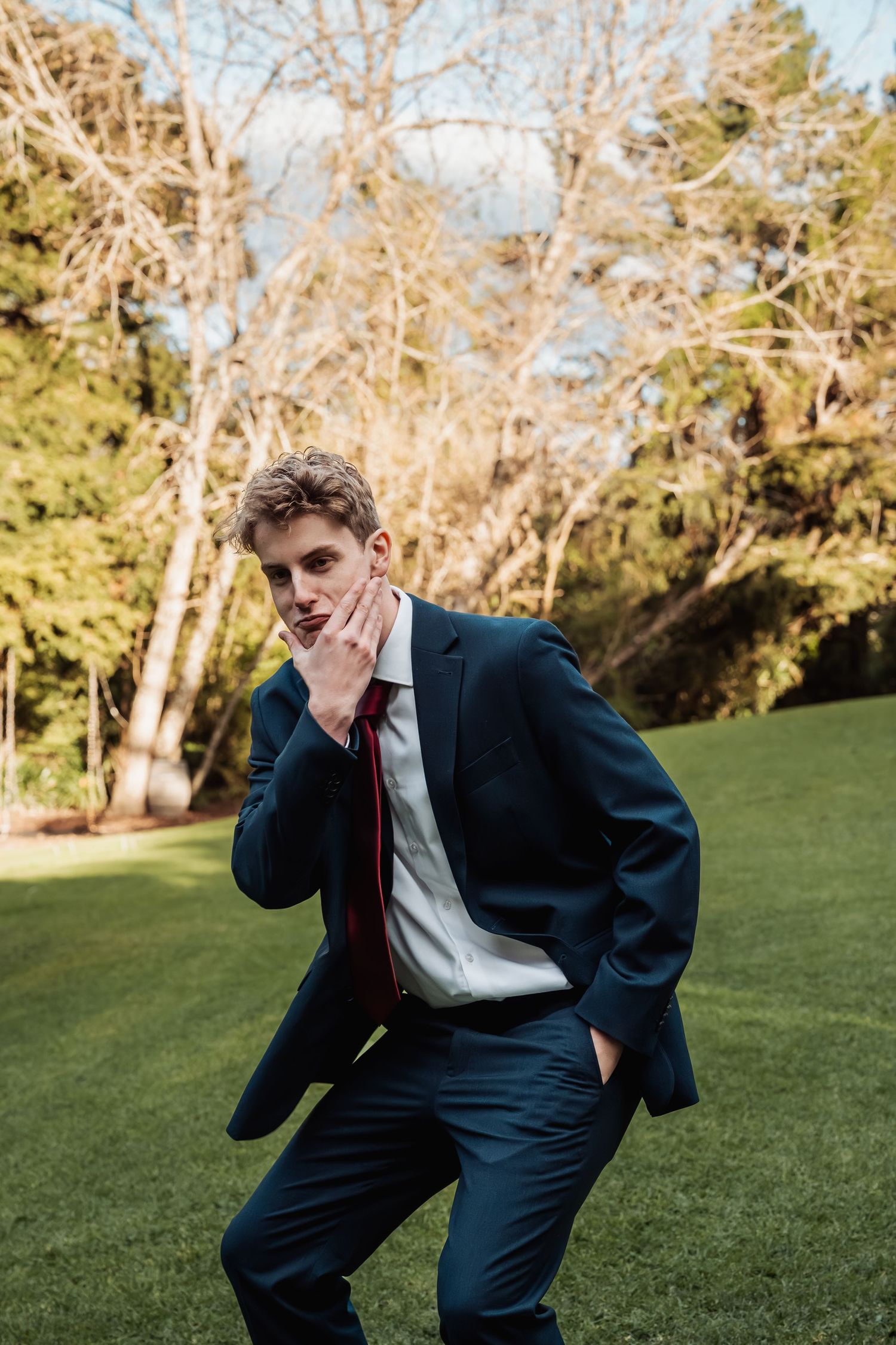 Young man in navy blue suit with red tie posing outdoors on green grass with trees in background.