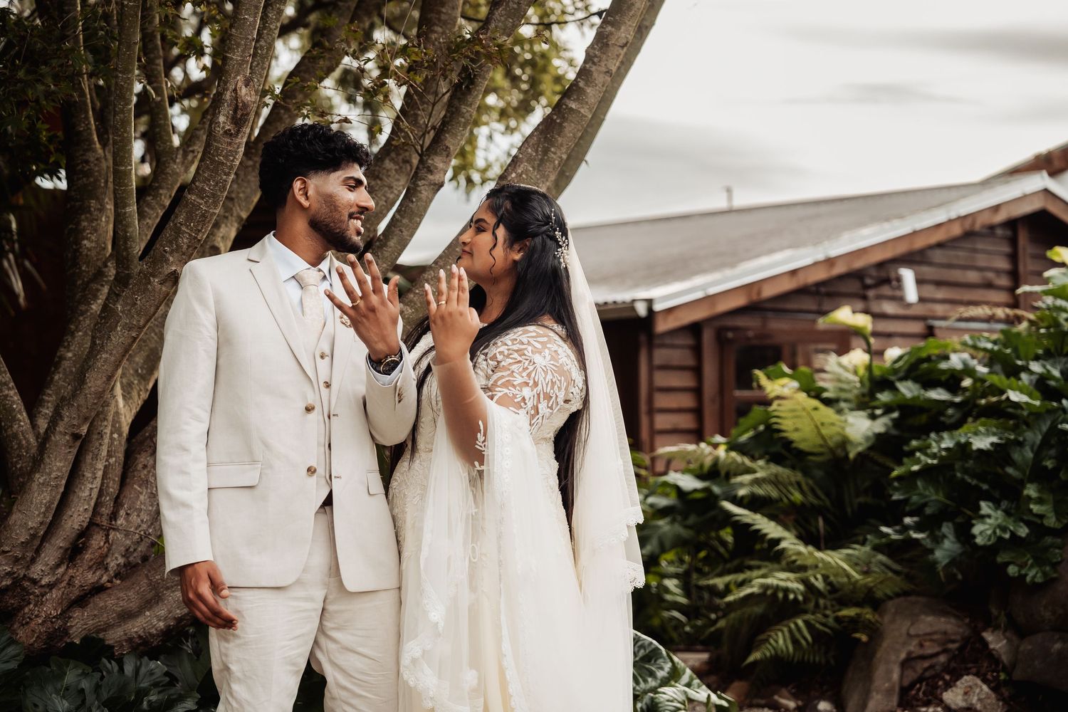 Bride and groom share a joyful moment together beneath large tree in lush garden setting.