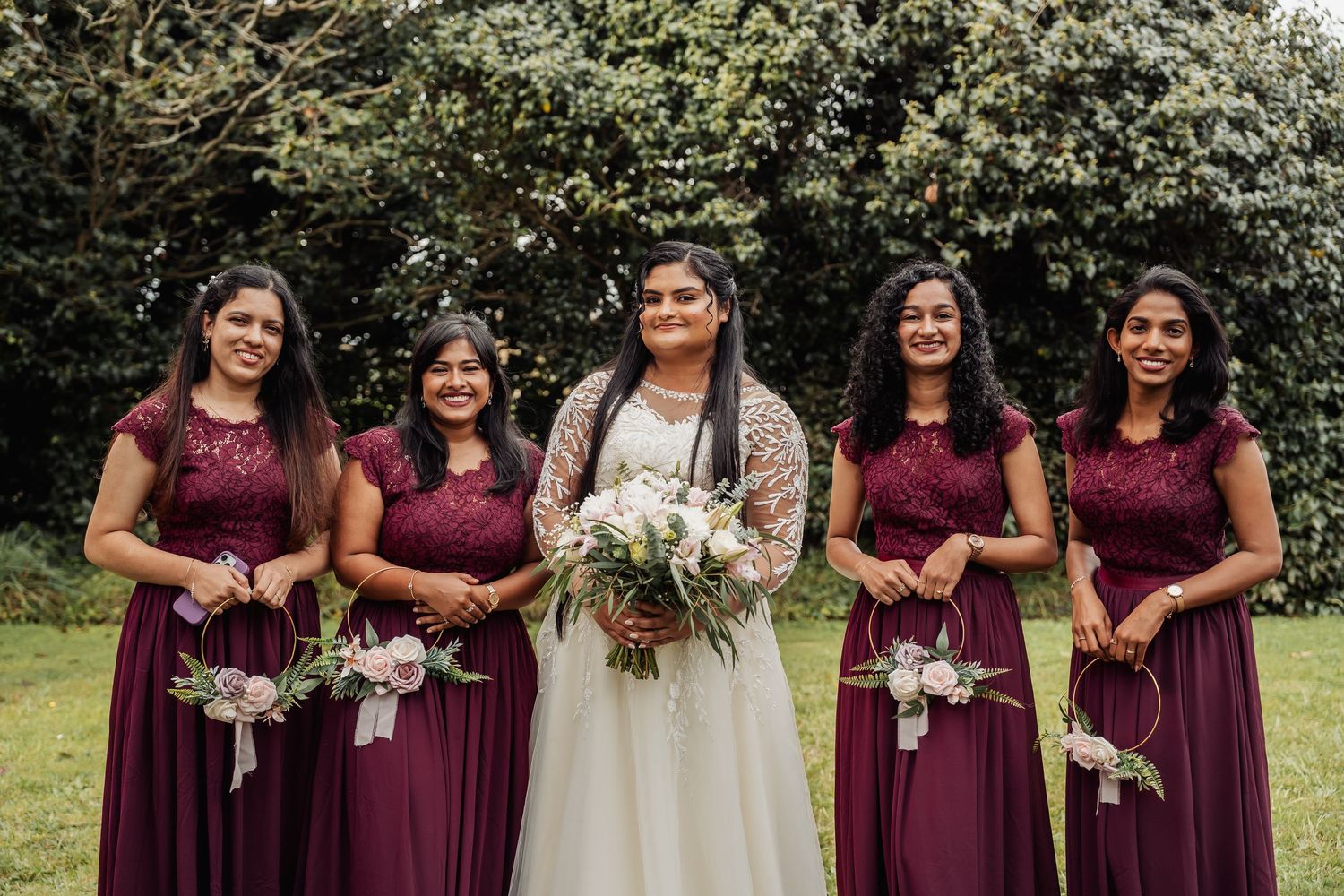 Bride in white gown with four bridesmaids in burgundy dresses holding bouquets outdoors.