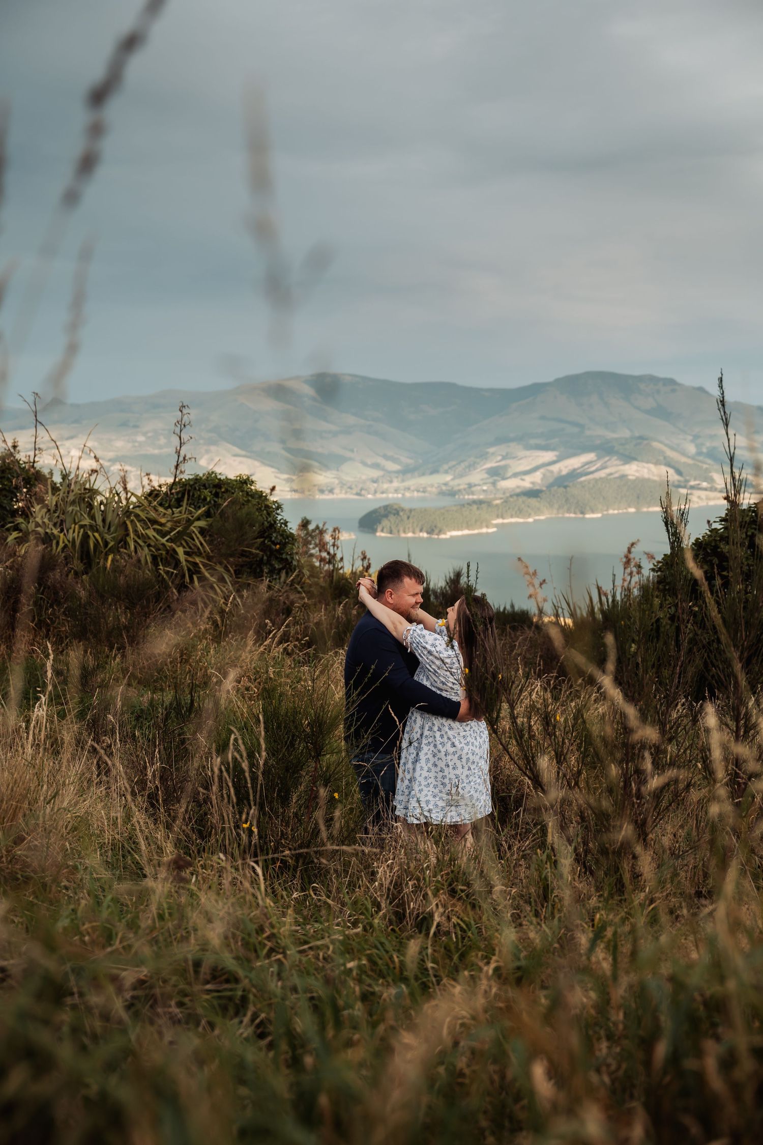 Couple embracing in tall grass field with scenic mountain and coastal backdrop under cloudy skies.
