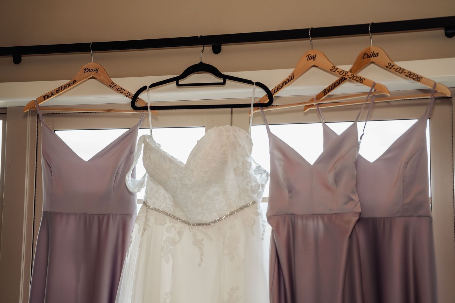 Wedding dress and bridesmaid gowns in dusty purple hanging on wooden hangers by a bright window.
