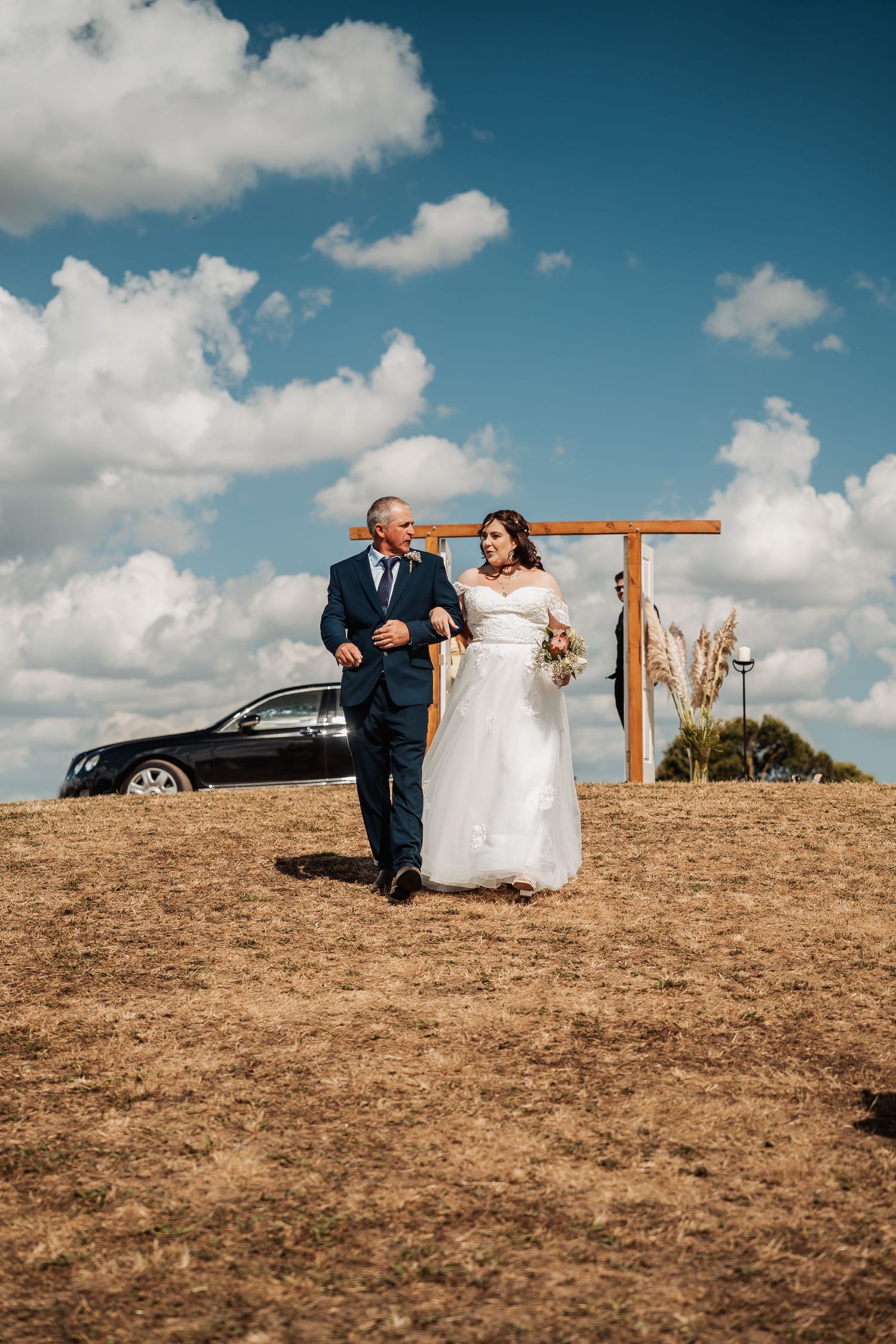 Bride in white gown walks with suited man past wooden arch under blue cloudy sky on dry grassy hilltop.
