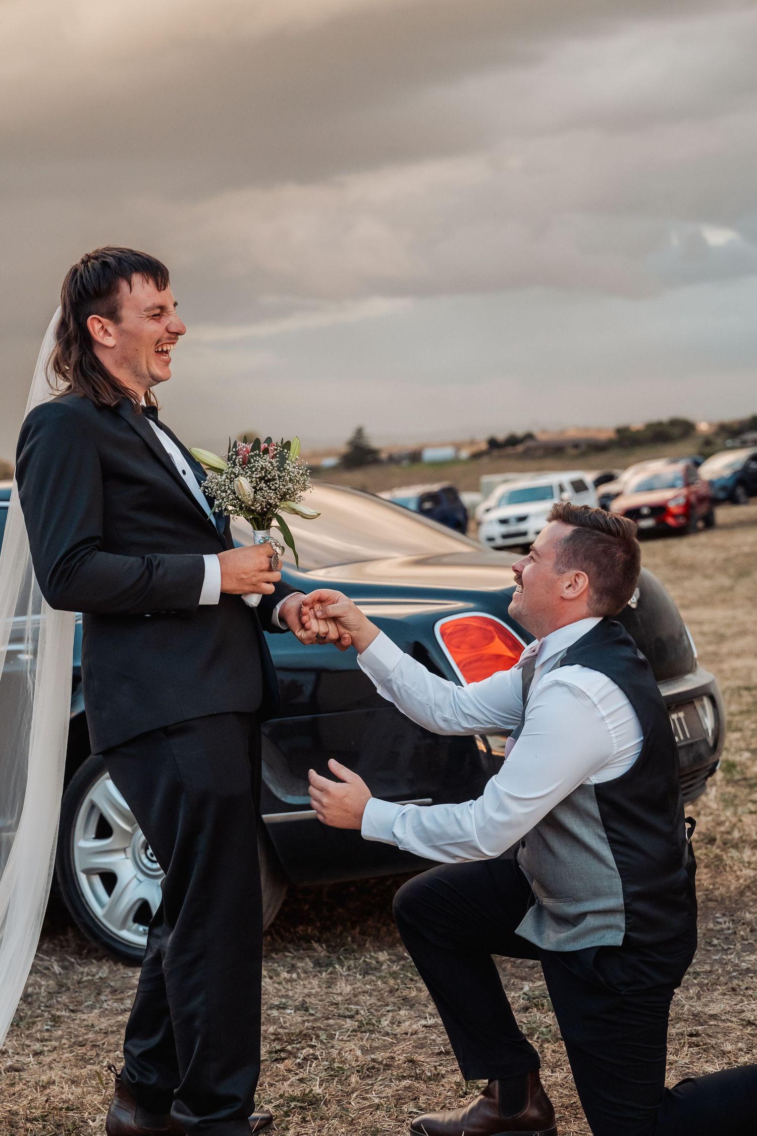 Groom laughing as groomsman kneels playfully before him in a wedding bouquet handoff near a black car.