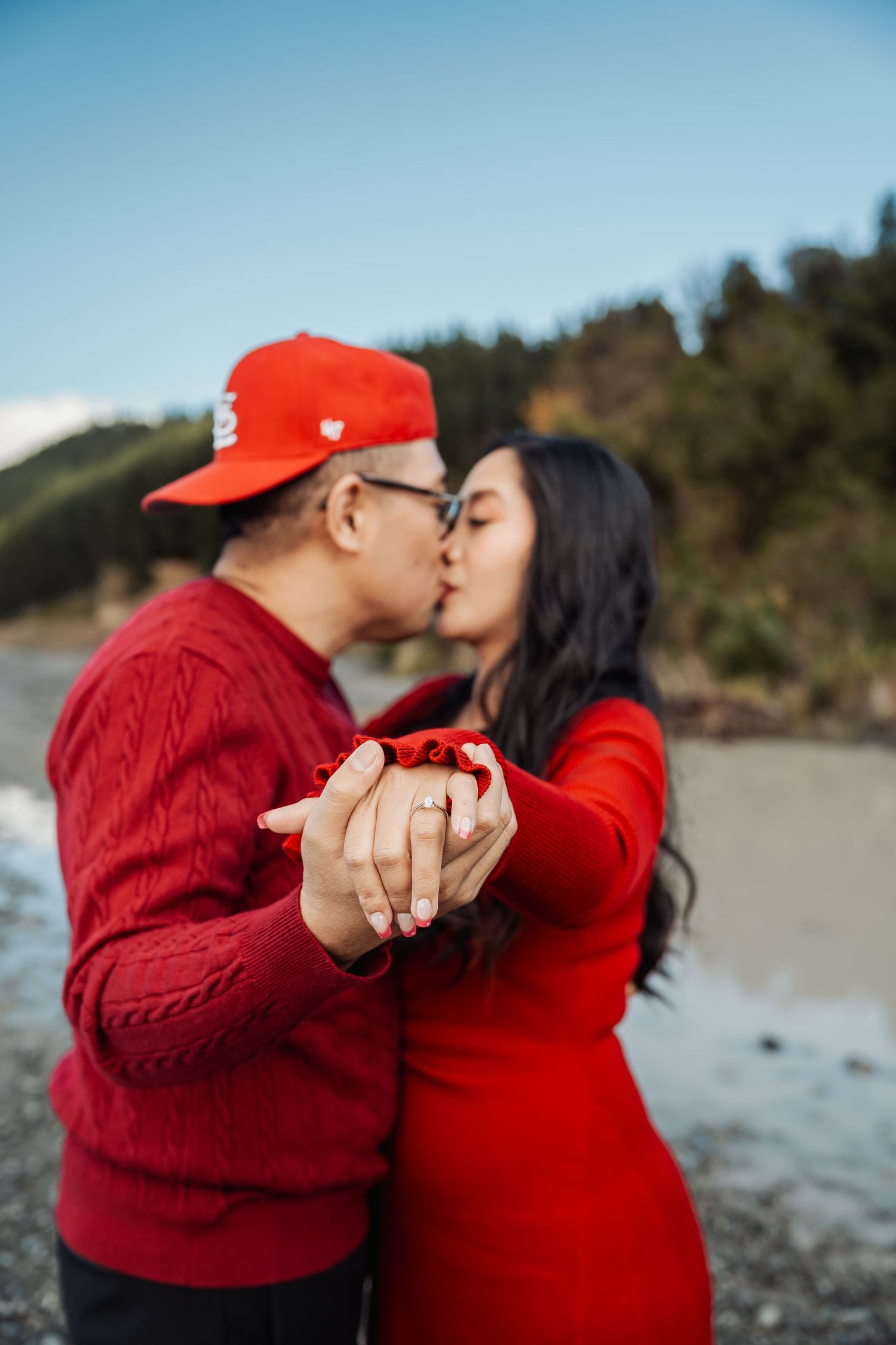 Engaged couple in red outfits kiss while displaying a diamond ring near a peaceful shoreline.