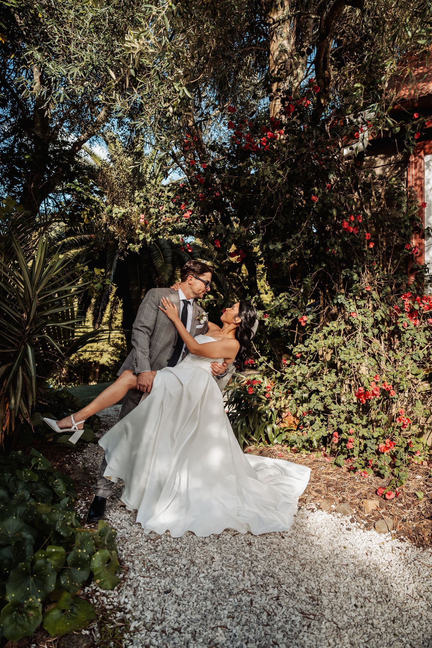 Groom dips bride for a kiss on garden path surrounded by vibrant red flowers.