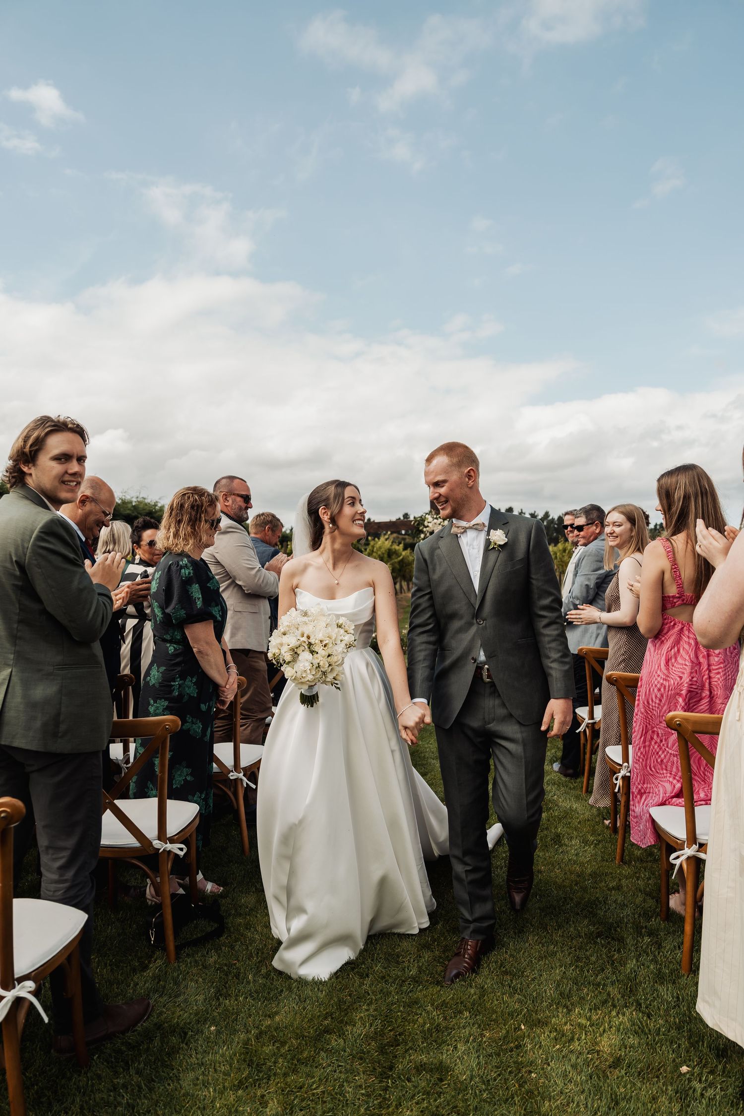 Happy couple walks down grass aisle as wedding guests celebrate at an outdoor vineyard ceremony.