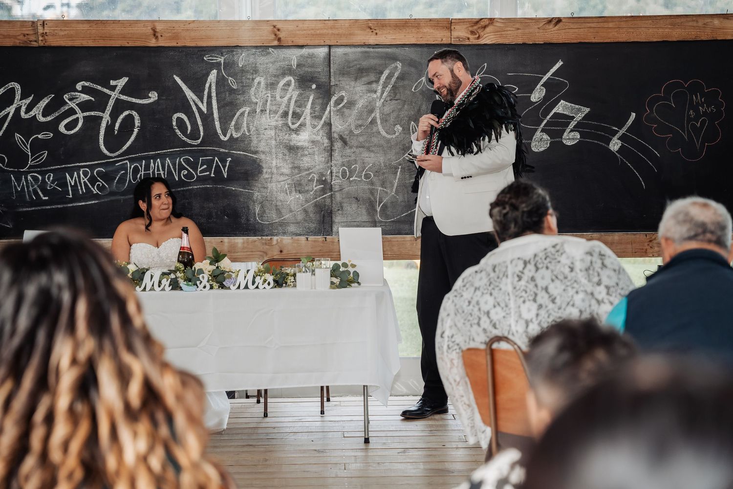 Groom gives a speech at wedding reception in front of a just married chalkboard sign.