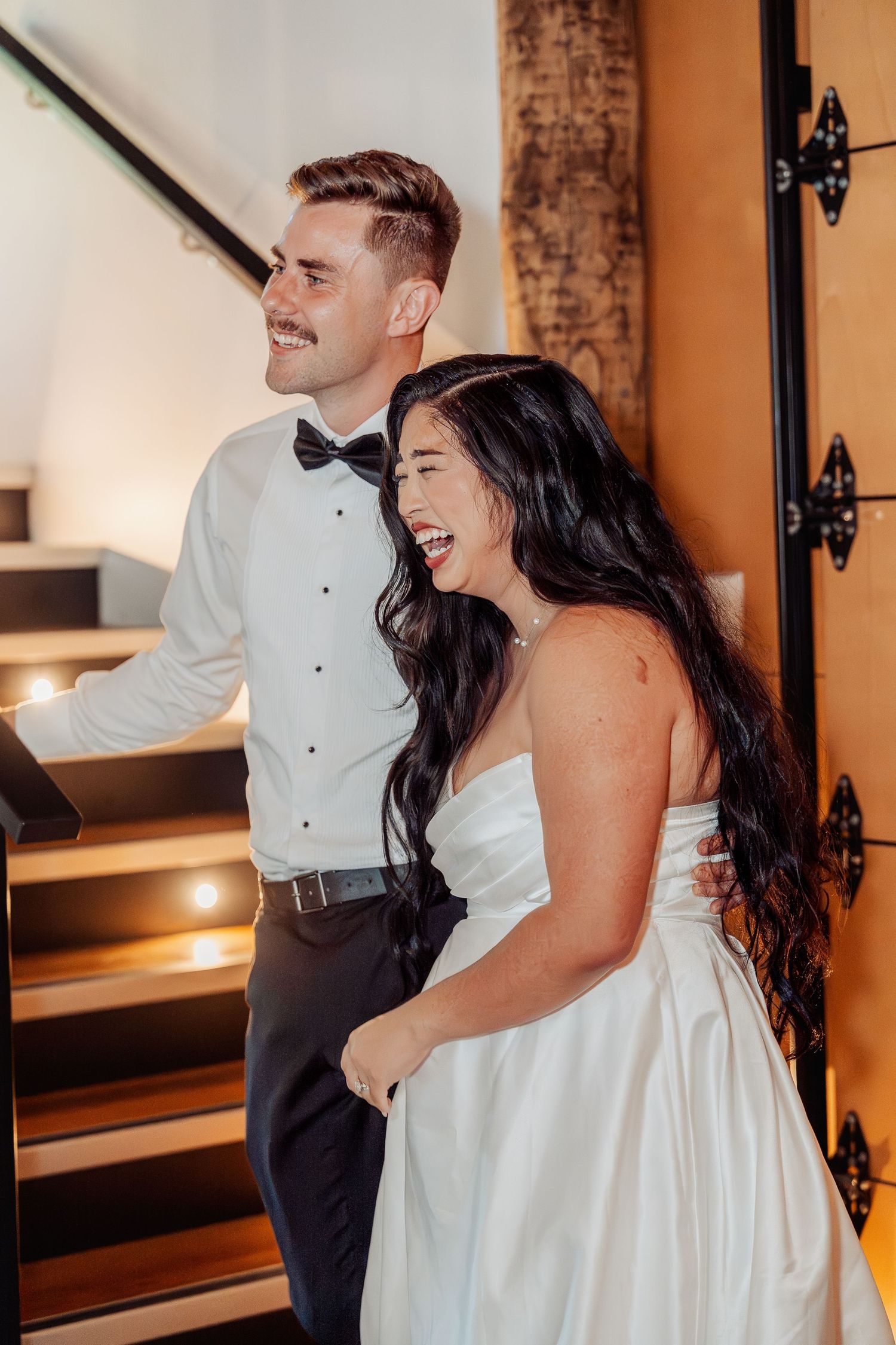 Laughing bride in strapless white gown and groom in bow tie walking up illuminated staircase together.