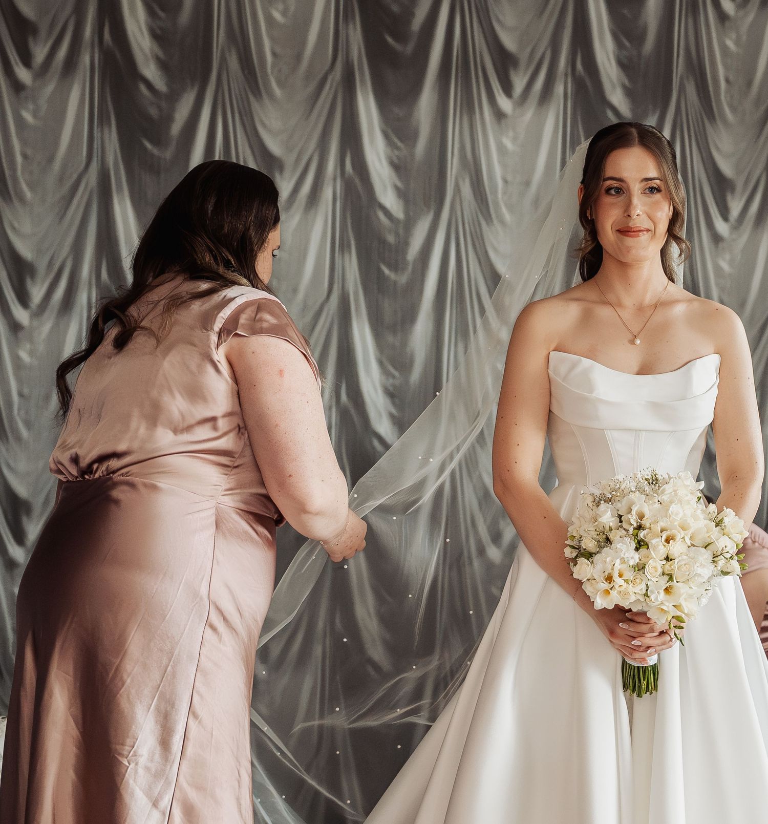 Bride in strapless white gown holds floral bouquet while bridesmaid in pink satin dress adjusts veil against draped backdrop.