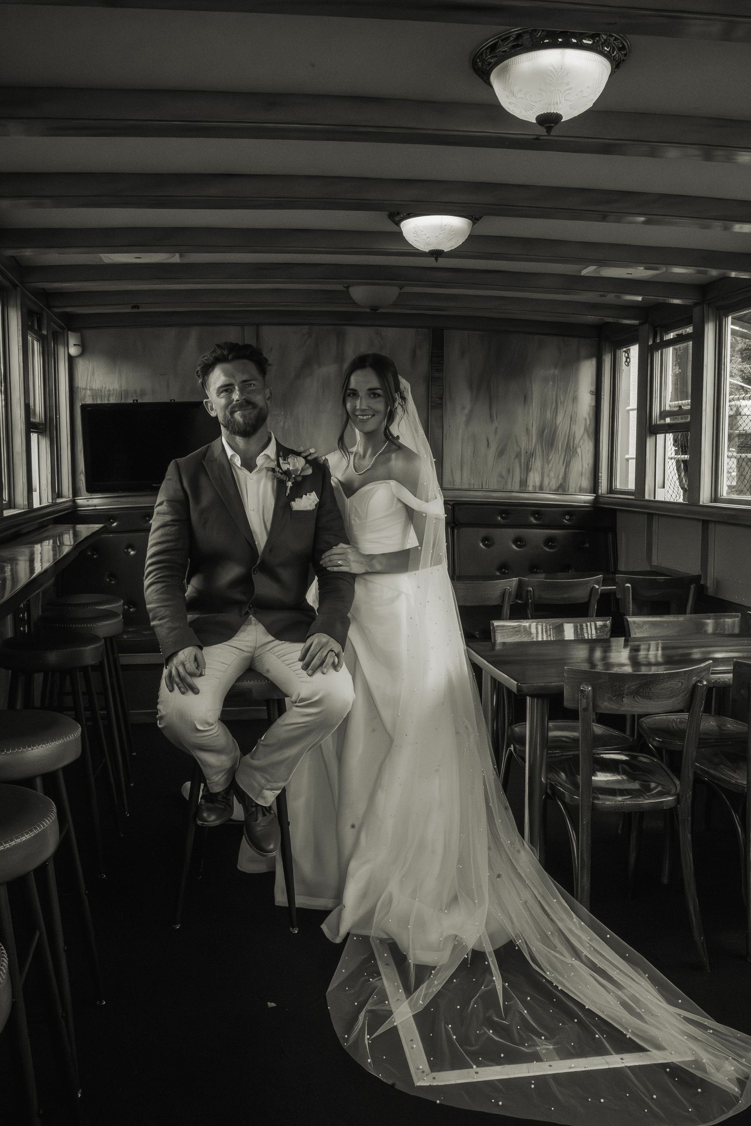 Black and white portrait of bride and groom posing elegantly inside vintage wooden boat interior.