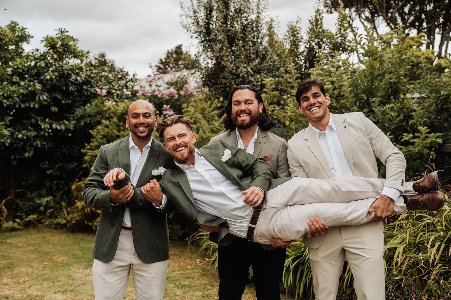 Four groomsmen in sage green and beige suits celebrate outdoors, one being carried playfully.