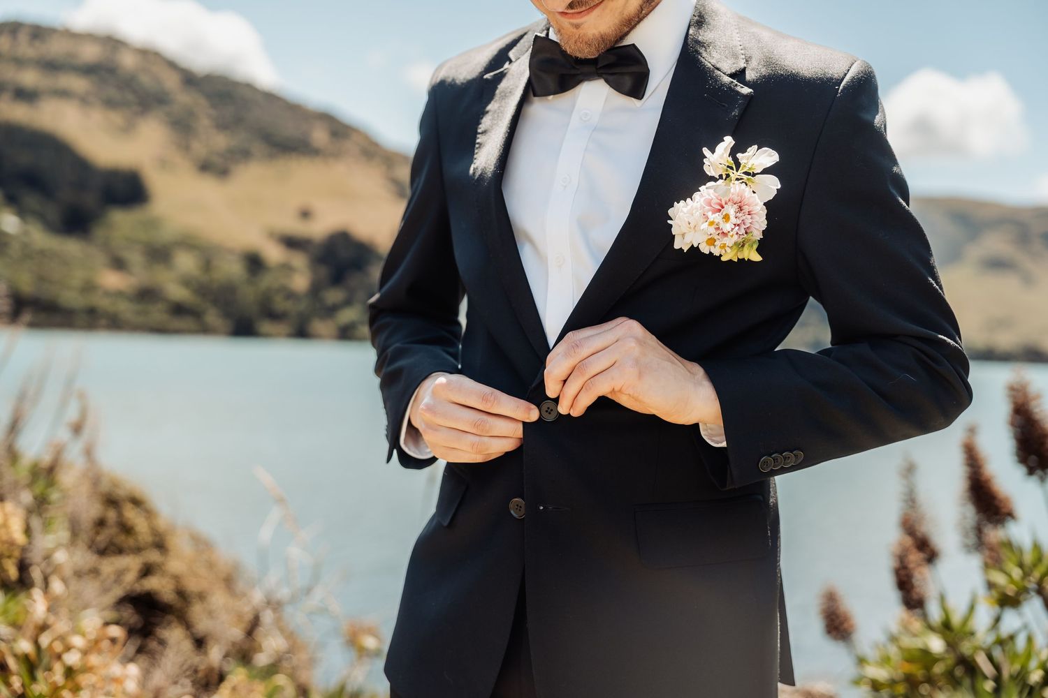Groom in navy tuxedo with bow tie and floral boutonniere buttoning jacket near lakeside venue.