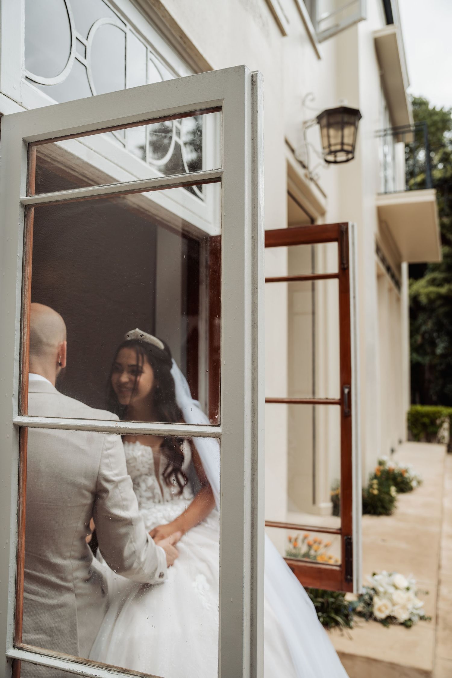 Bride and groom share tender moment viewed through white-framed window of wedding venue.