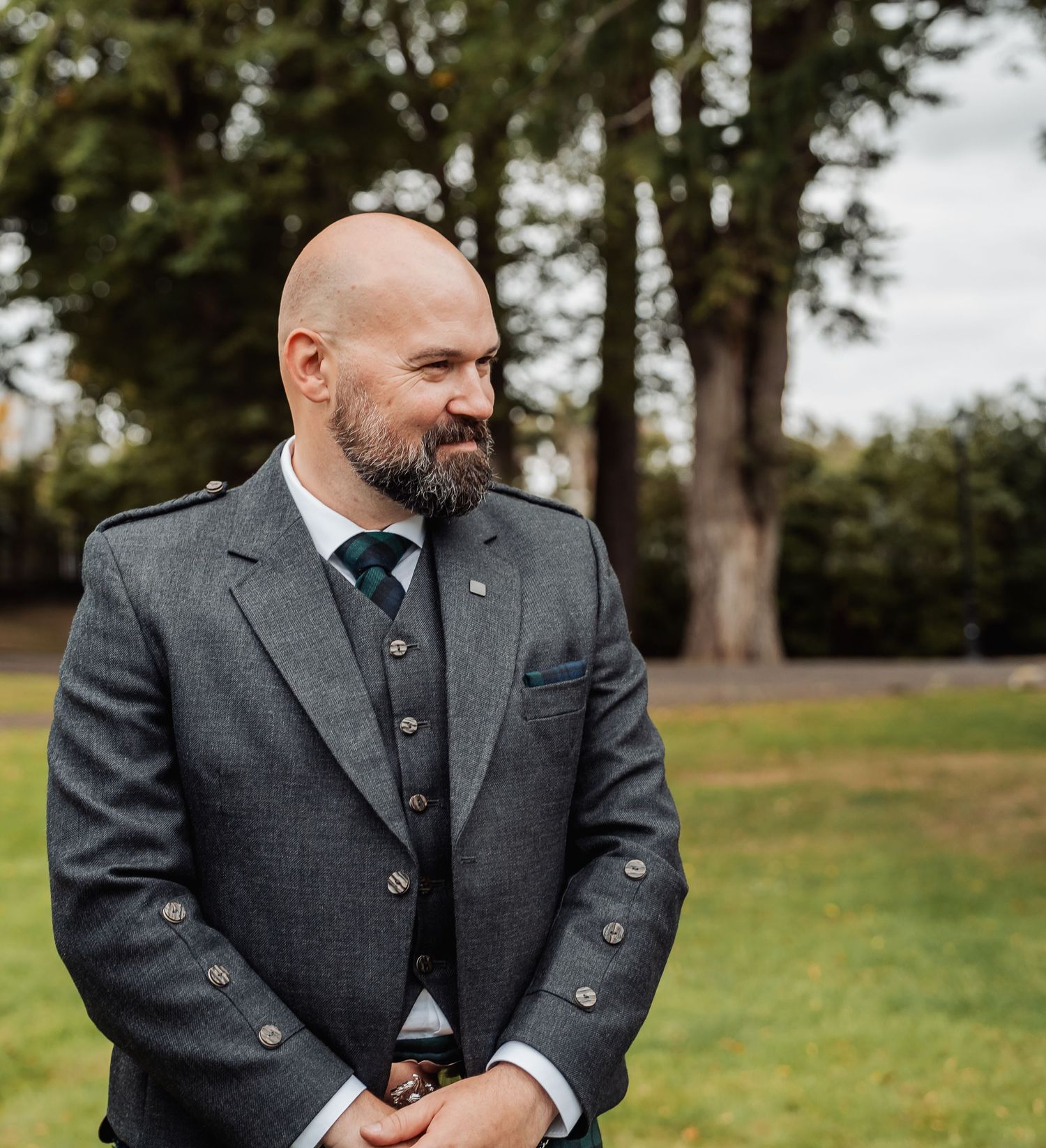 Bald groom with beard wearing grey Scottish tweed jacket and teal tie stands outdoors at wedding.