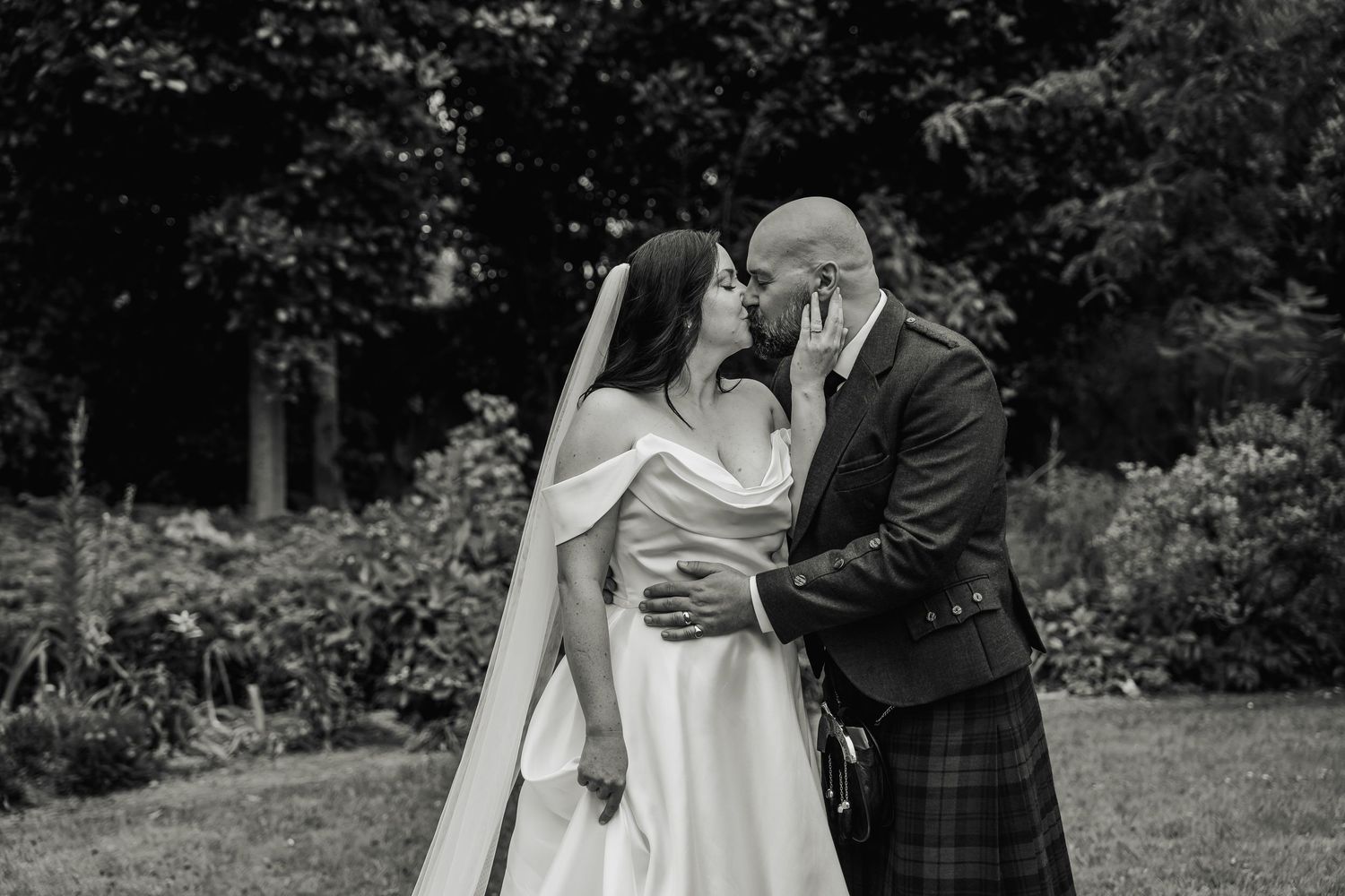 A black and white photo of a bride and groom sharing a romantic kiss in a lush garden.