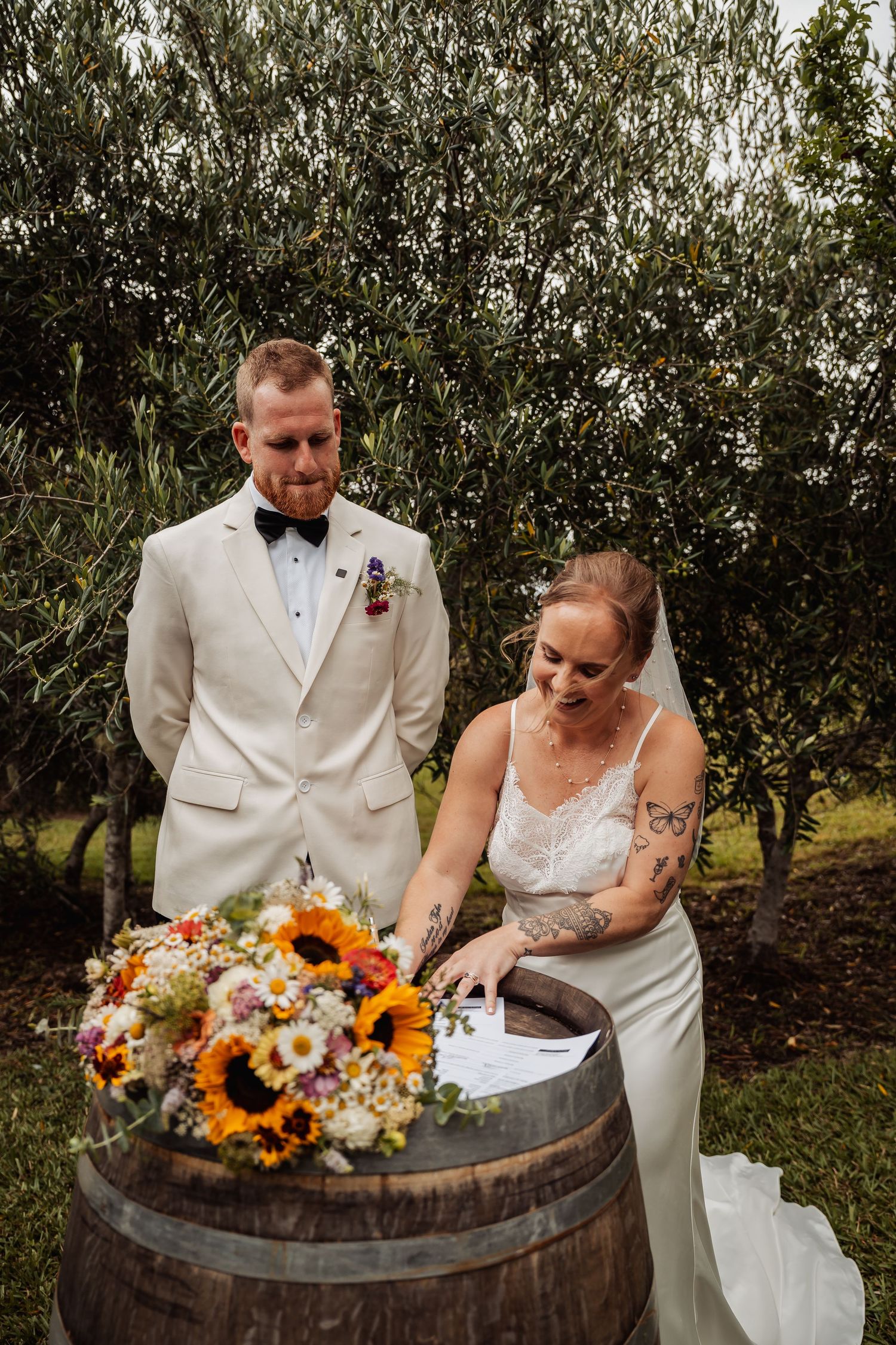 Bride and groom signing wedding register on a barrel with colorful sunflower bouquet outdoors.