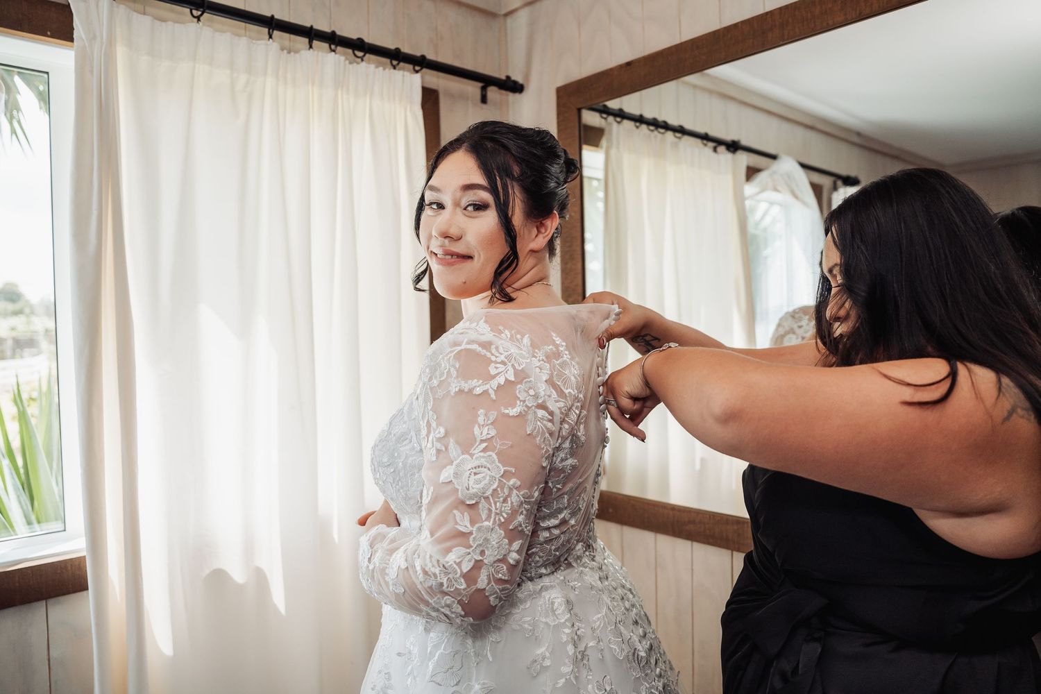 Someone helps adjust a delicate white lace wedding dress with long sleeves in a sunlit room with white curtains.