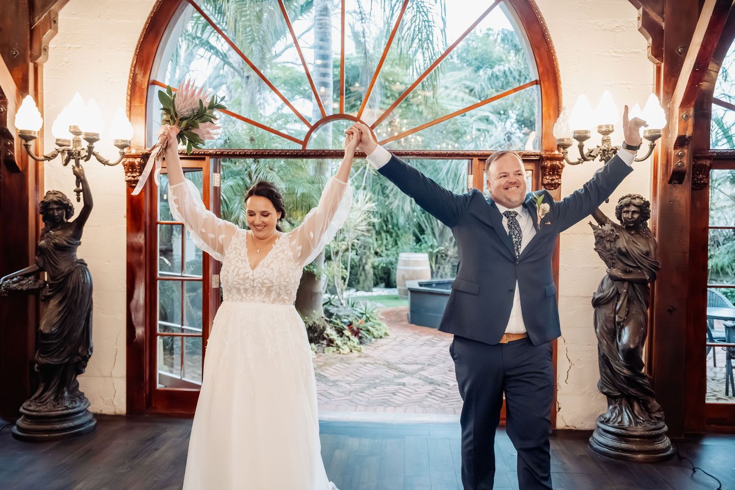 Wedding couple celebrates with arms raised in an ornate archway surrounded by tropical foliage.