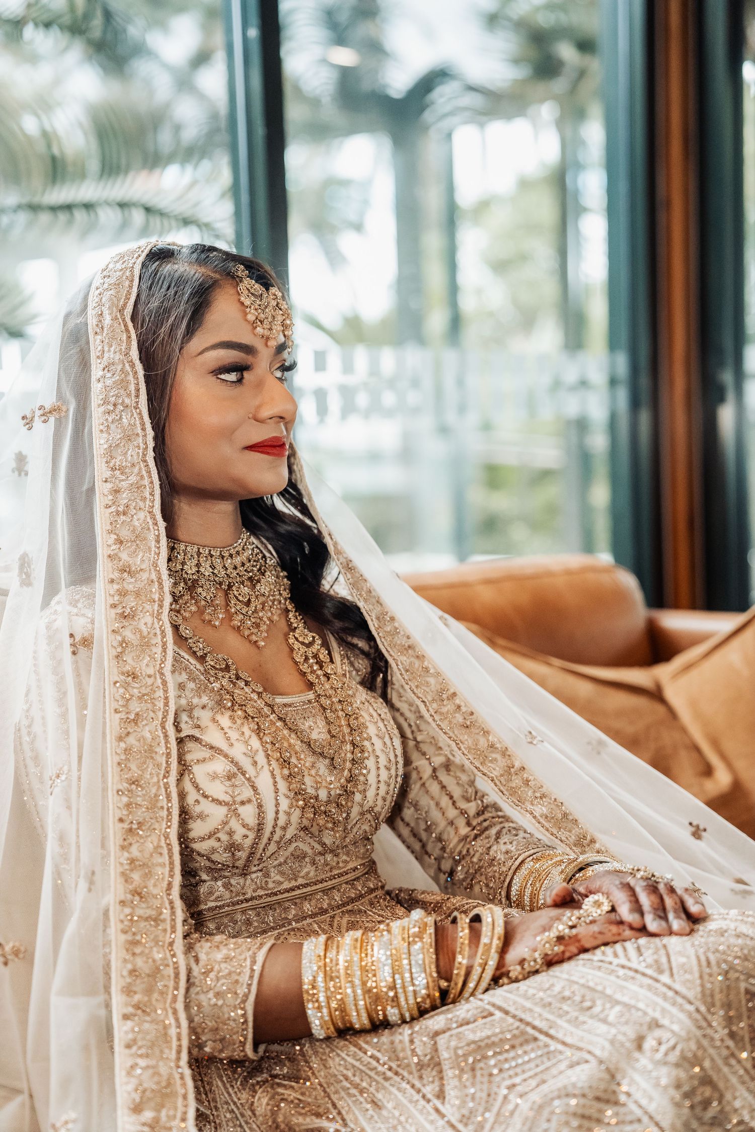 A bride in an ornate gold embroidered lehenga and delicate veil poses elegantly on a brown leather couch.
