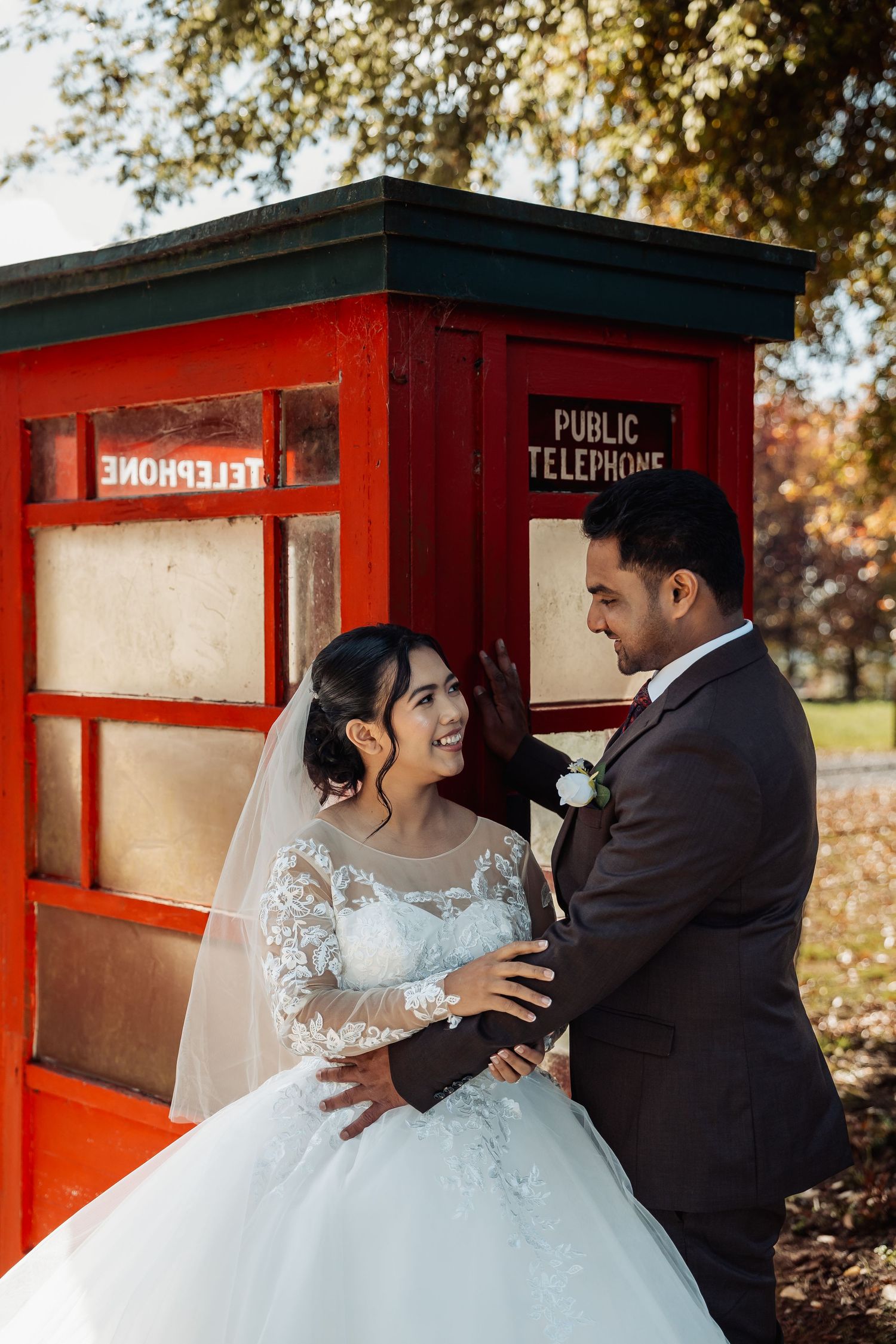 Bride and groom sharing a joyful moment beside a vintage red public telephone booth outdoors.