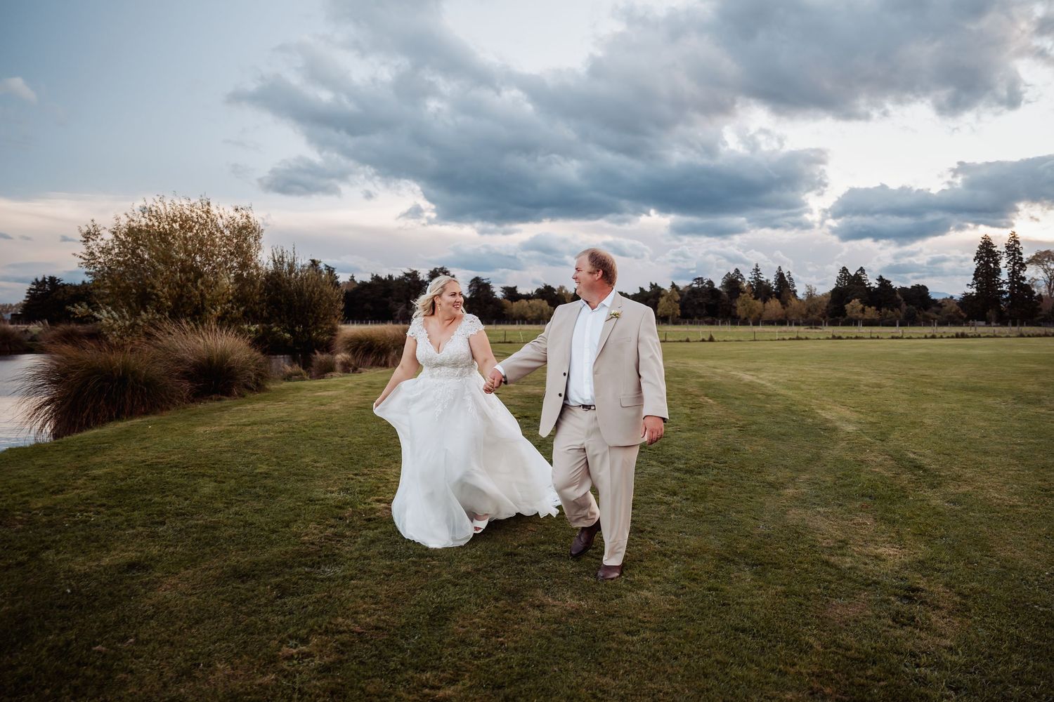 Bride and groom walk hand in hand across green lawn at sunset with dramatic cloudy sky.