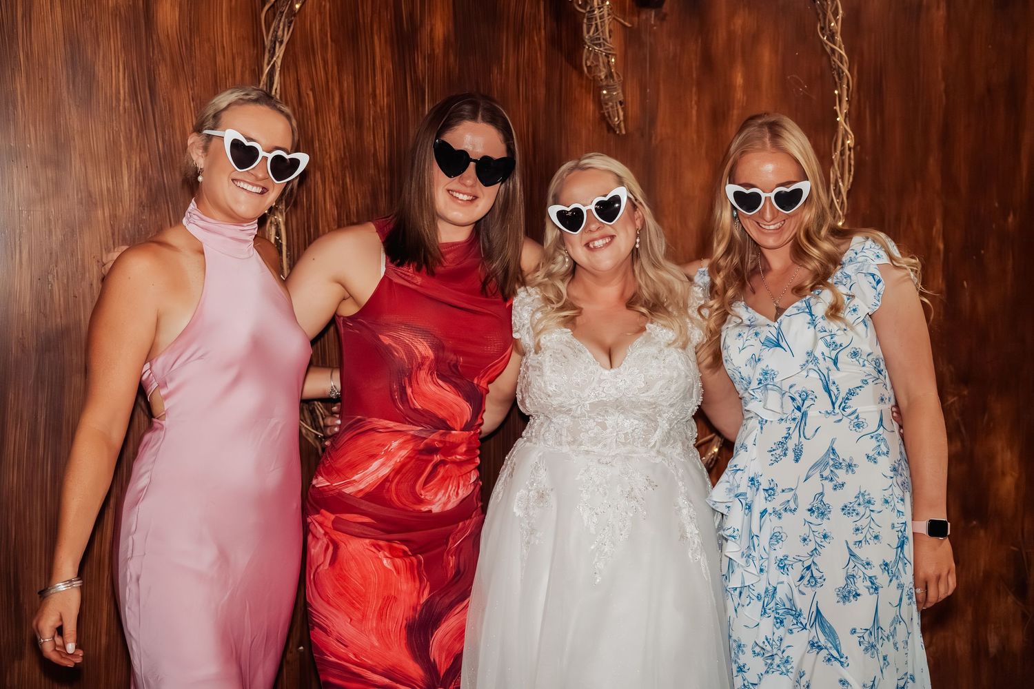 Bride in white gown smiling with three guests in pink, red, and blue dresses wearing fun sunglasses.