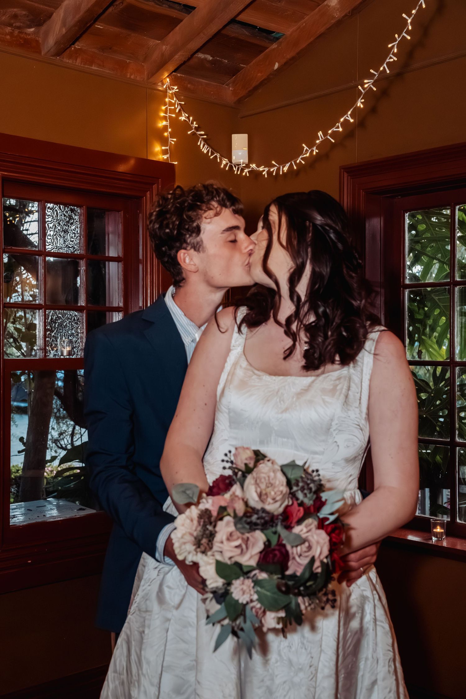 Newlyweds share a kiss holding bridal bouquet with fairy lights in background.