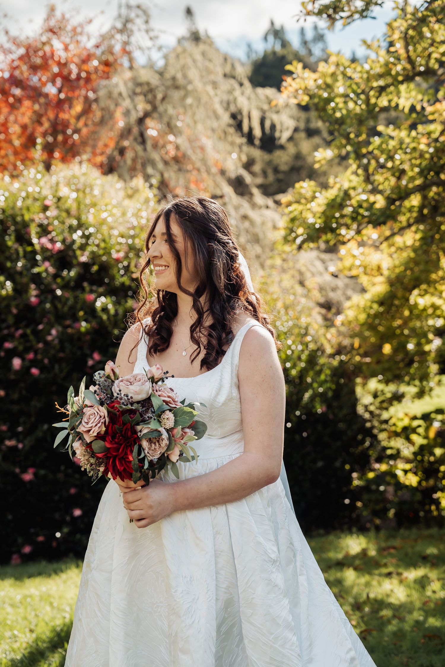 Smiling bride in white gown holding red and pink bouquet in sunny garden setting.