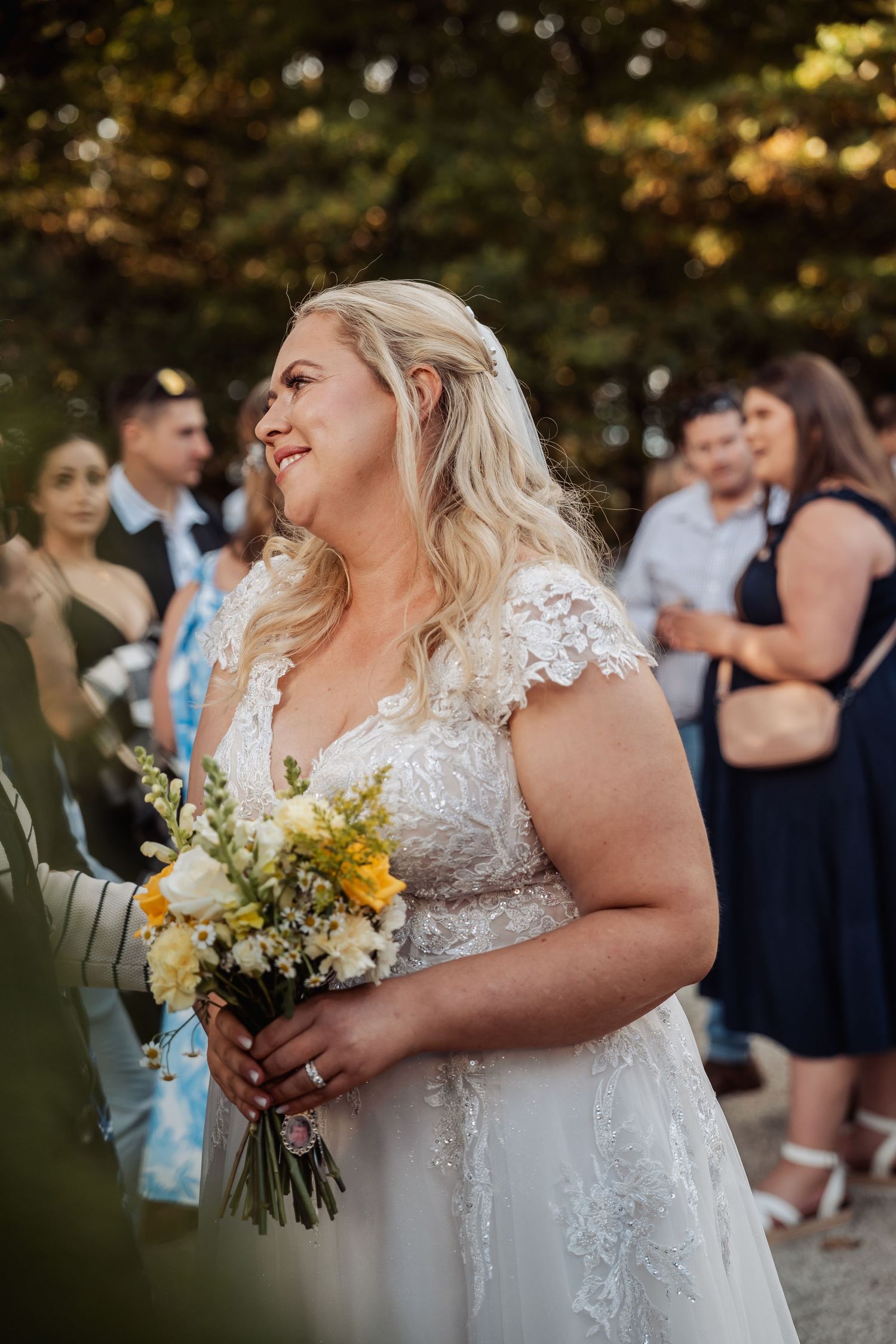 Smiling bride in lace wedding gown holds yellow and white bouquet during outdoor wedding celebration.
